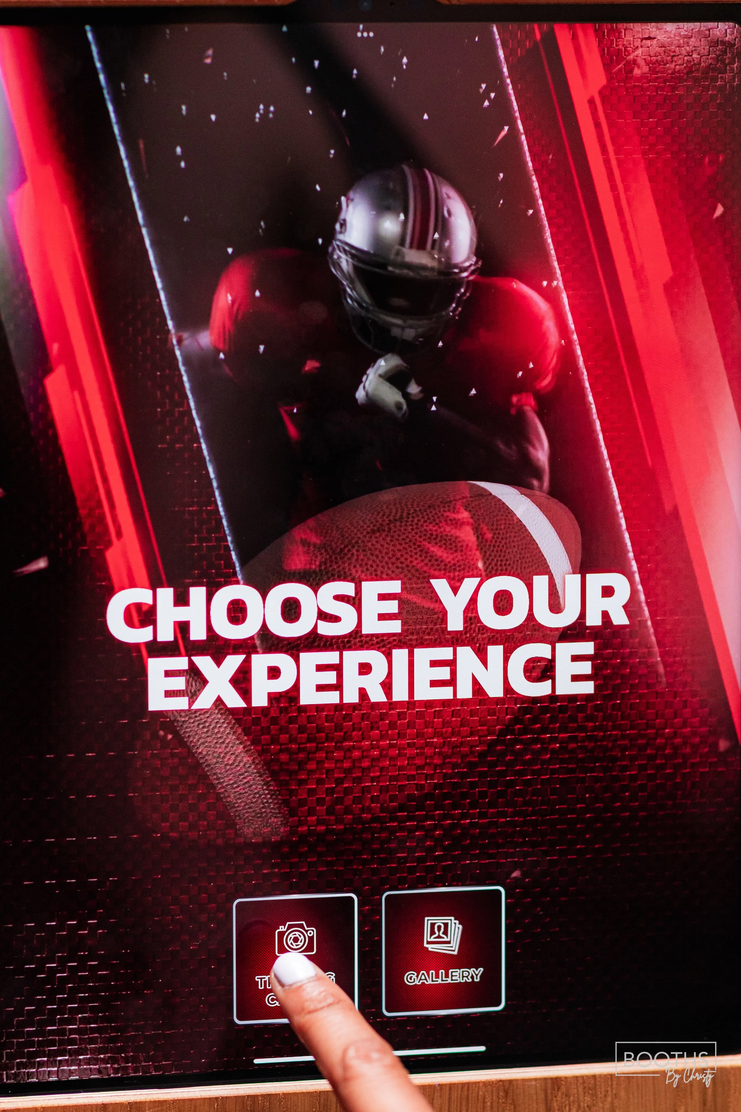 Digital display with a football player in helmet and uniform, with the text "Choose Your Experience"; buttons labeled "Take a Photo" and "Gallery" with a person's finger pointing at the photos option.