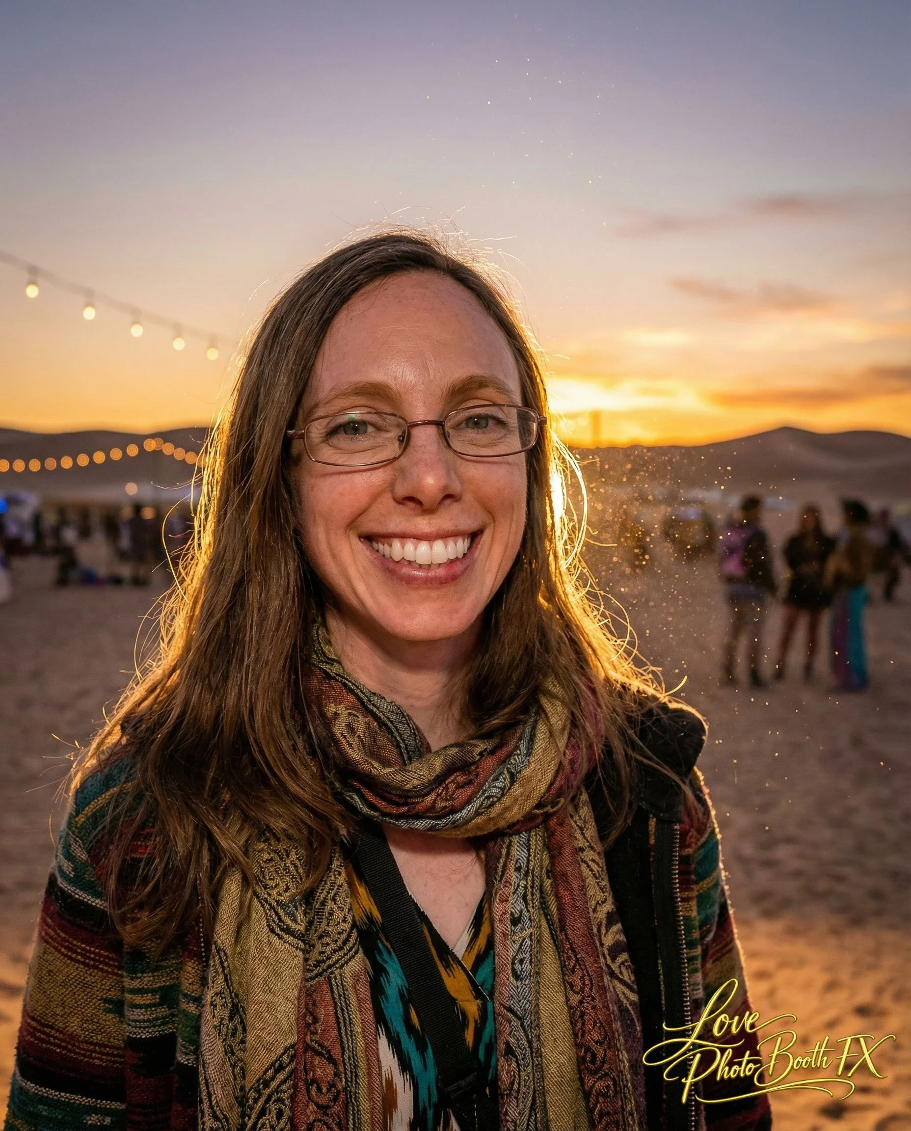 A smiling woman with glasses and a scarf, standing outdoors at sunset with a string of lights and people in the background.