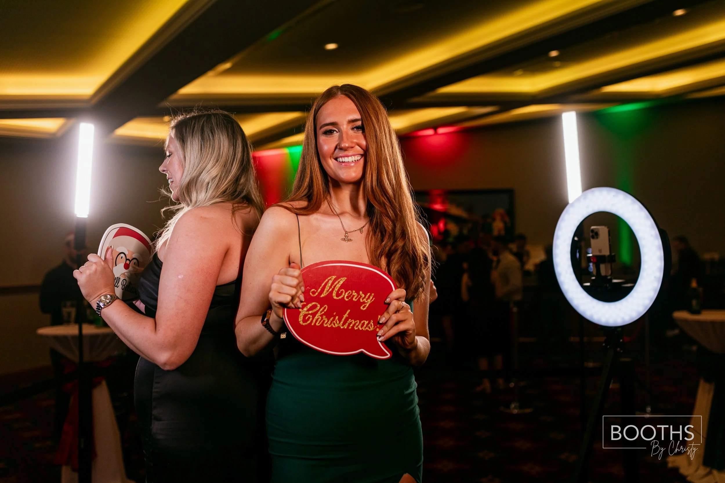 Two women at a holiday party, one facing forward smiling and holding a 'Merry Christmas' speech bubble, the other with her back turned holding a Santa Claus photo booth prop, standing in a decorated room with people and a ring light in the background