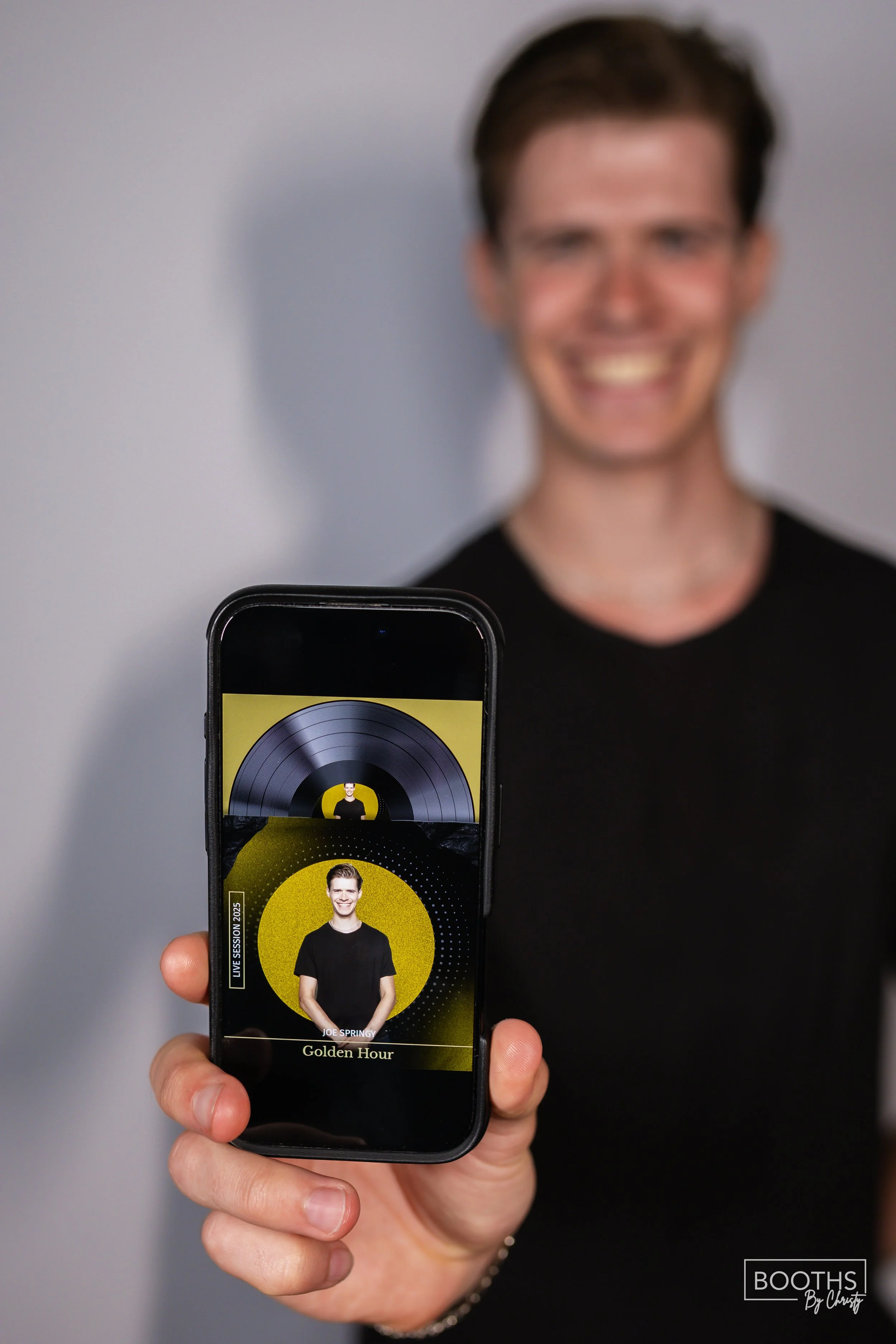 A person holding a phone displaying a music or event app with a photo of a smiling man in front of a yellow background, labeled 'Joe Springy' and 'Golden Hour', with a blurred background of the person's face.