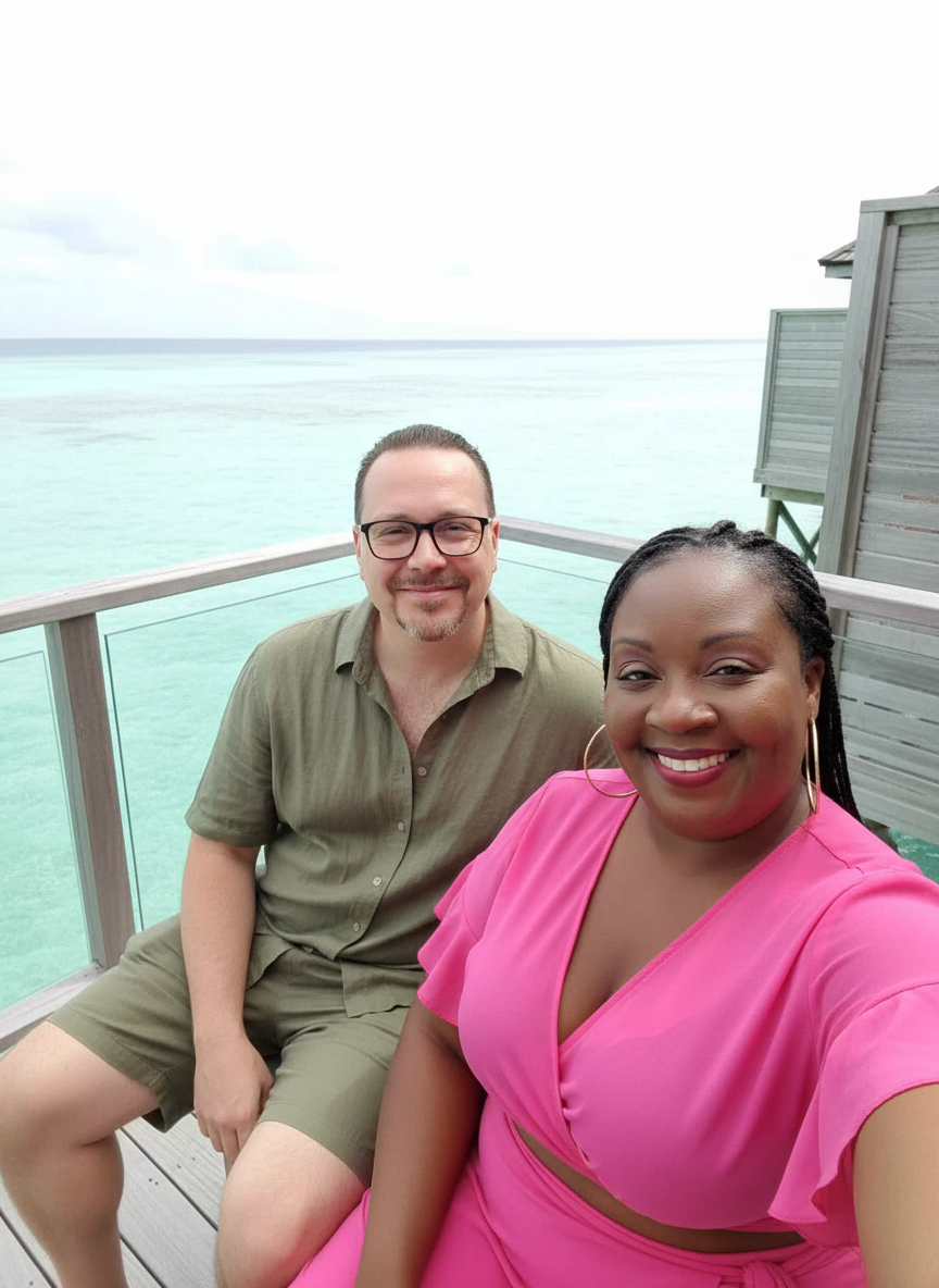 A smiling couple taking a selfie on a balcony overlooking turquoise water and a cloudy sky.