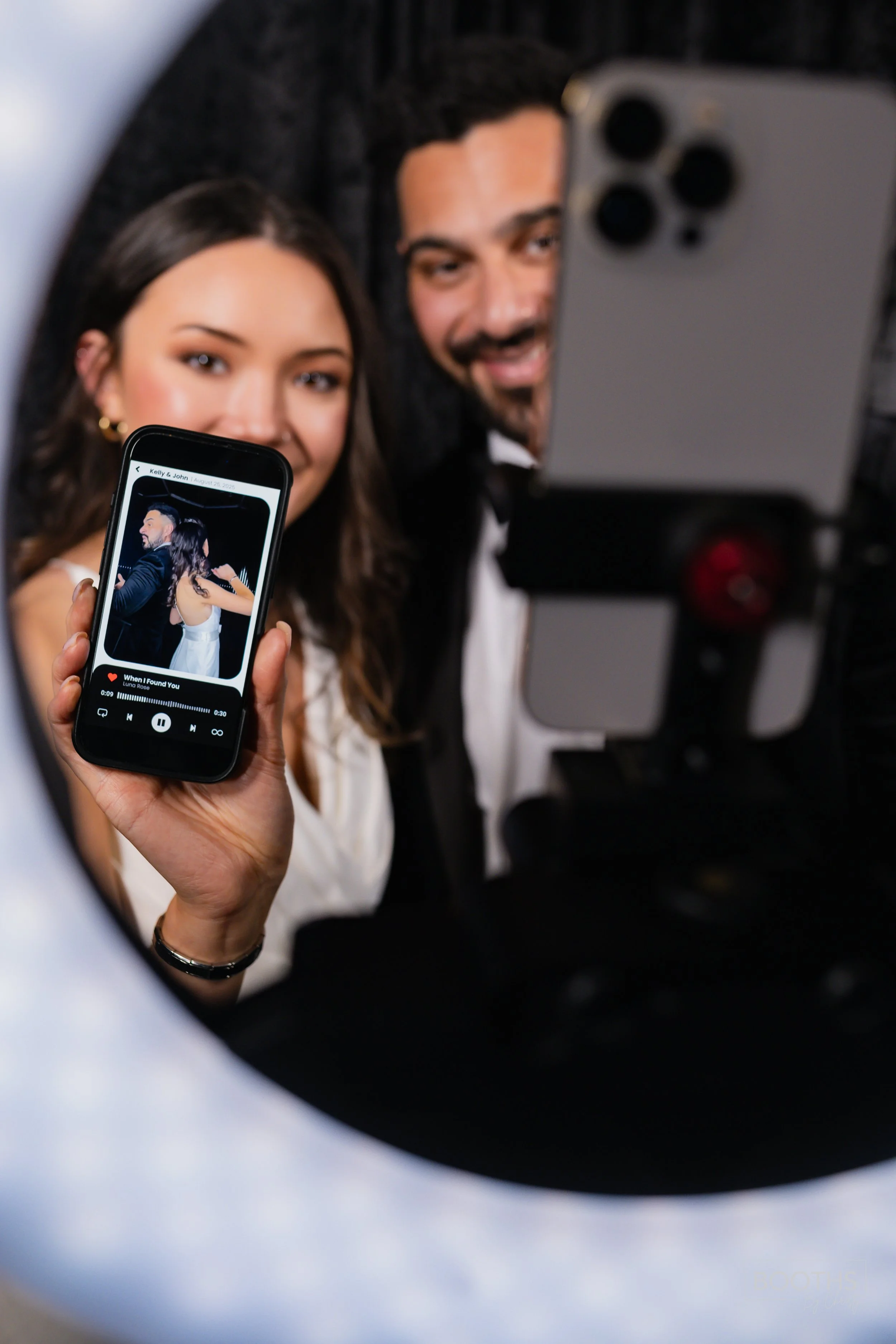 A couple taking a selfie while holding a phone displaying a music playlist; they are dressed in formal attire, likely at an event or celebration.