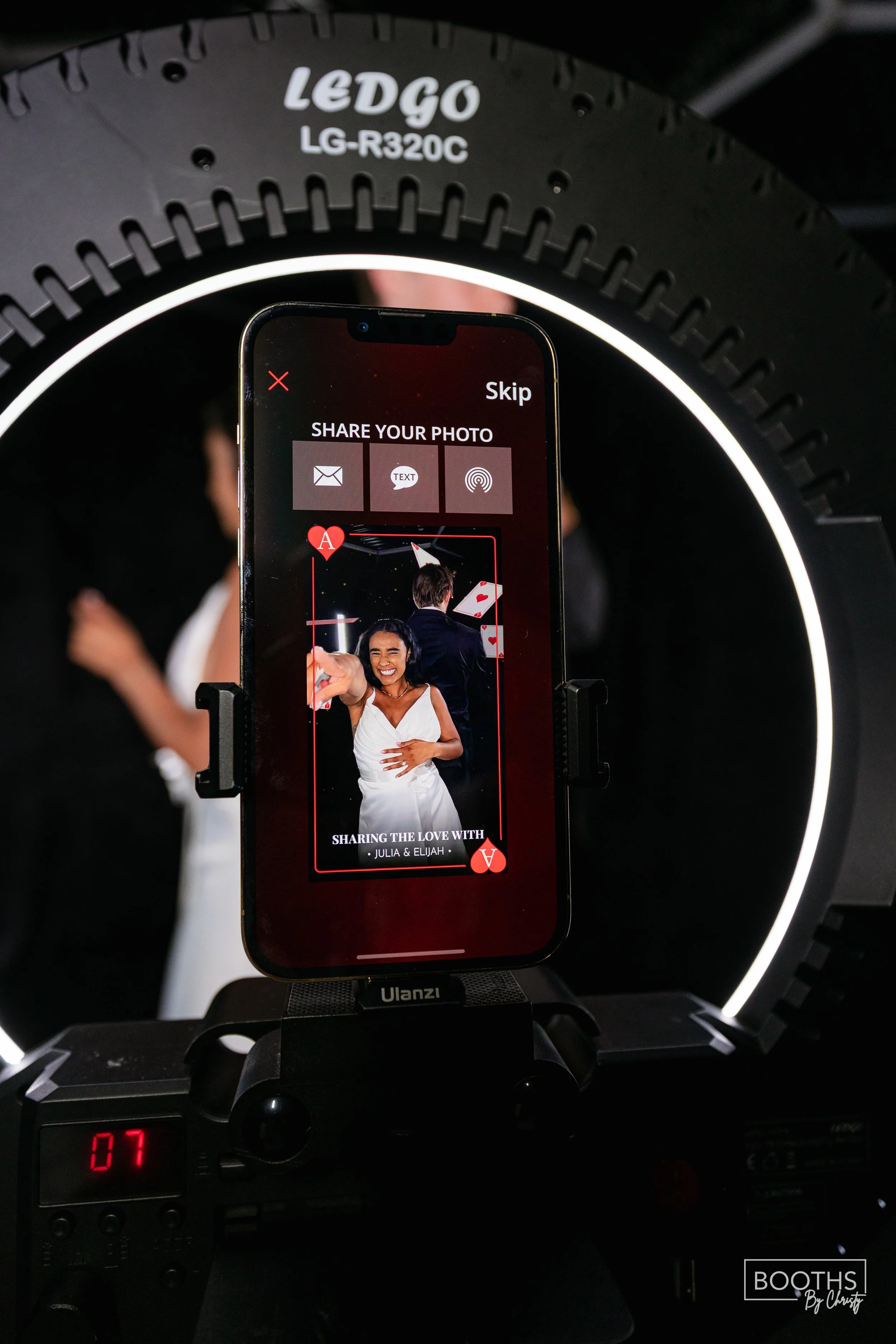 A smartphone mounted on a camera rig displays a photo-sharing app with a picture of a smiling woman in a white dress at a wedding, with a blurred couple in the background. The app offers sharing options including email, text, and a scanning feature,