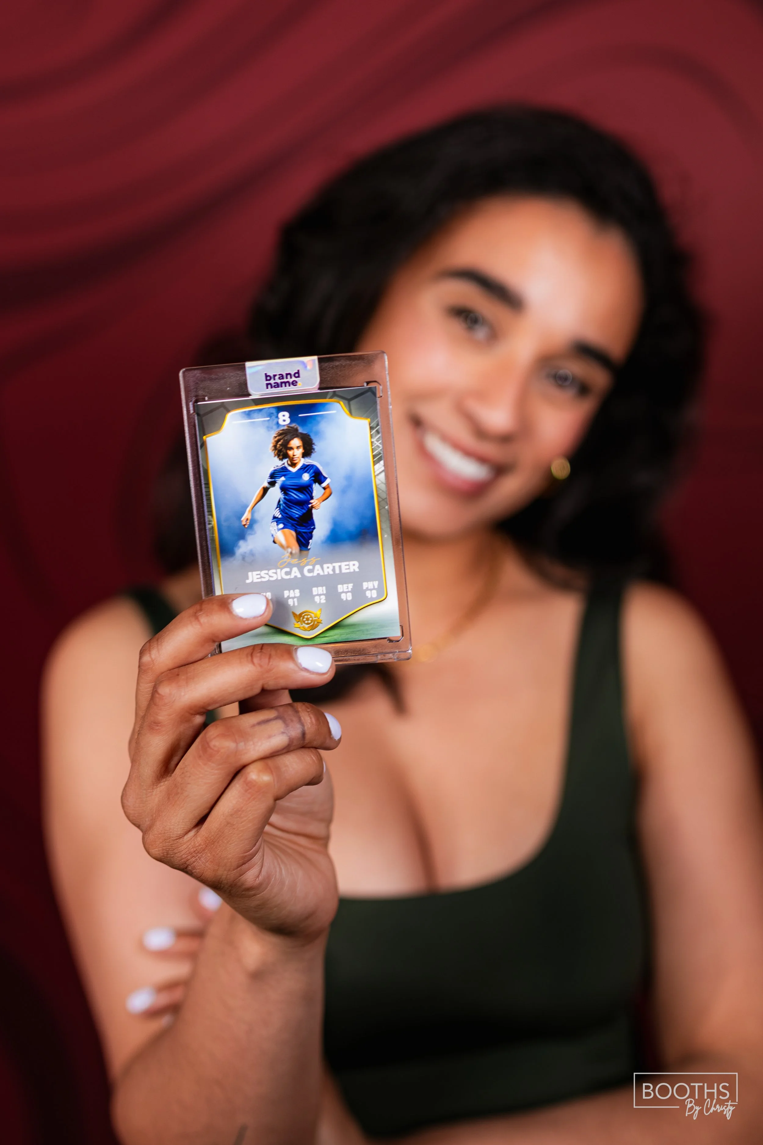 Woman holding a custom soccer player trading card featuring Jessica Carter with a blurred smiley face and a red wall background.