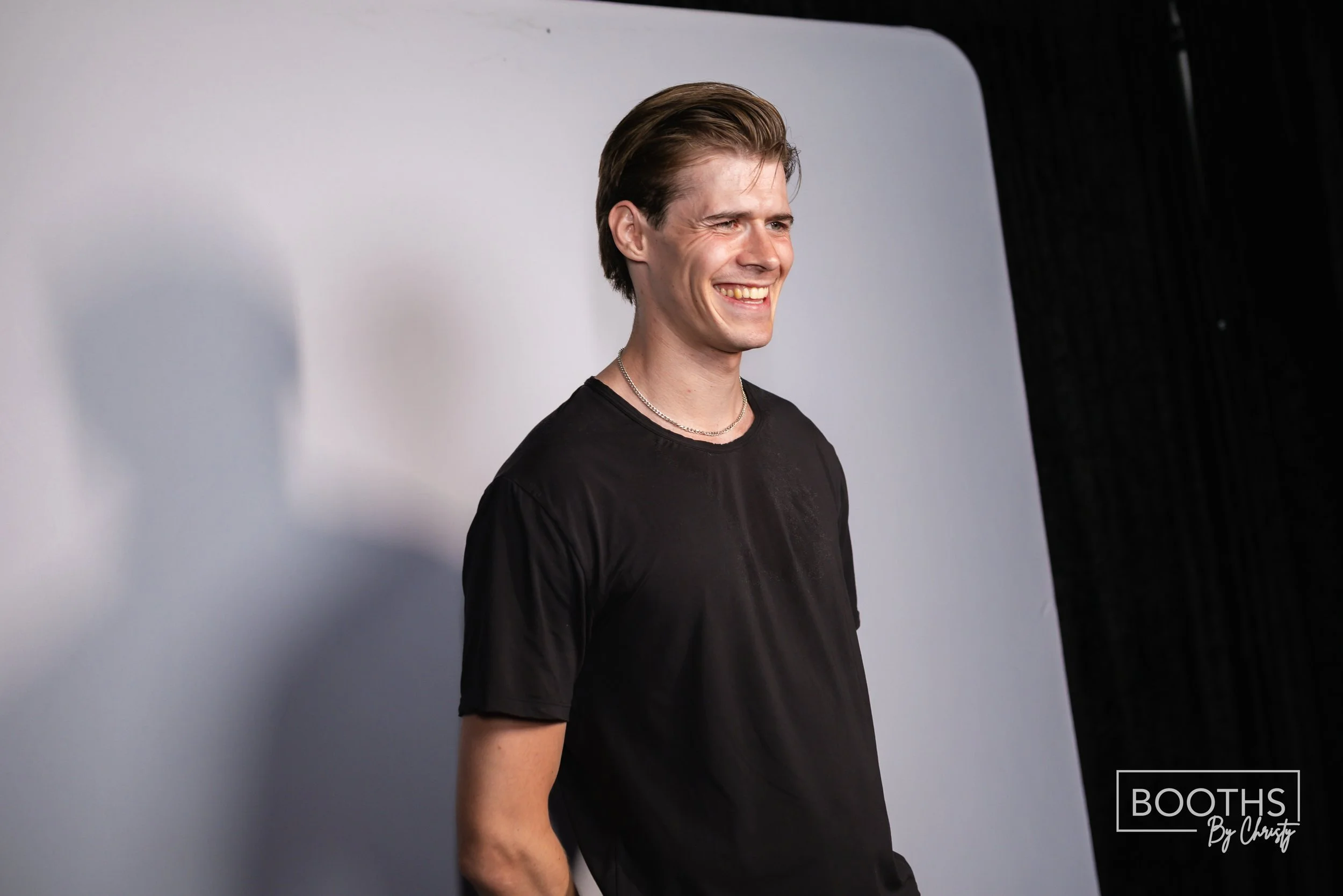 A young man with brown hair, wearing a black t-shirt and silver necklaces, smiling and standing against a white and black background.