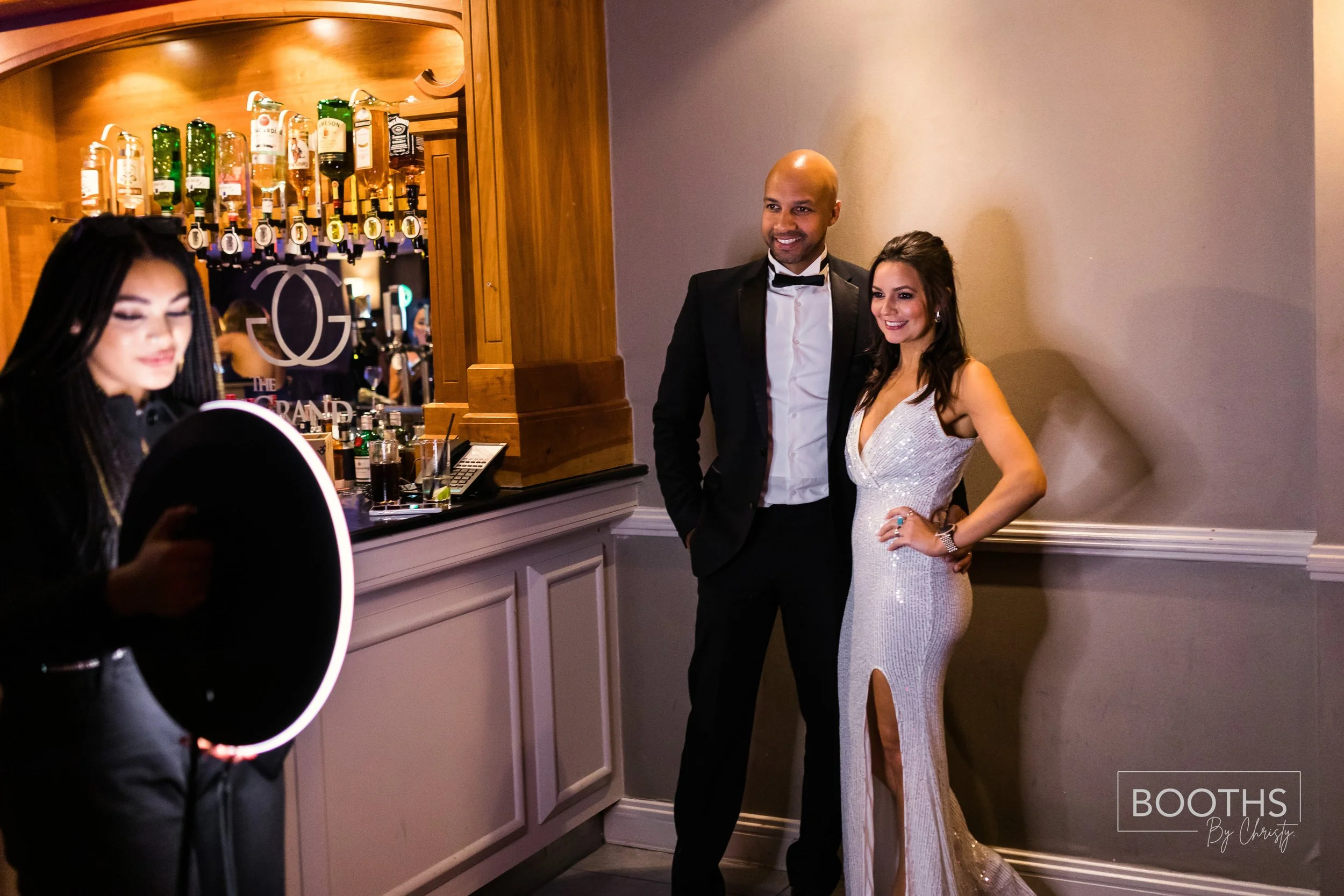 A man in a tuxedo and a woman in a white, silver, sequined gown posing together at a formal event, with a bartender in the foreground and a bar in the background.