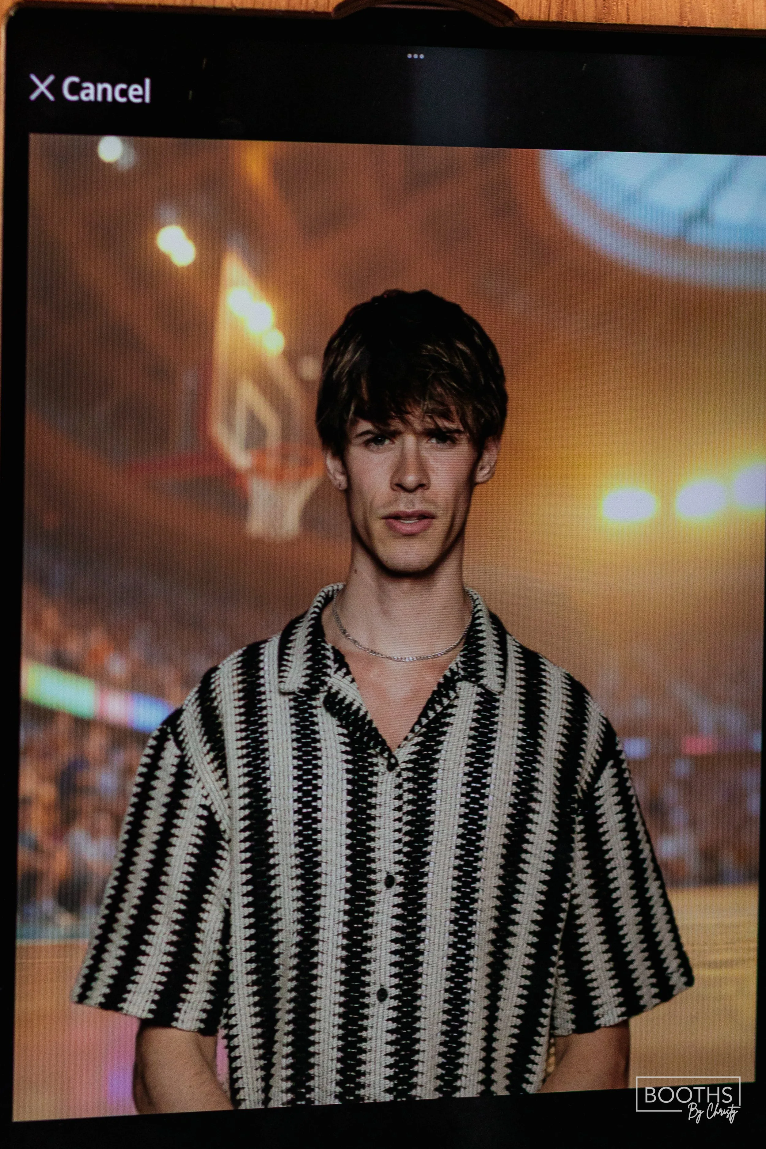 A young man with dark hair wearing a black and white patterned shirt and a silver chain, standing on a basketball court at sunset with a blurred crowd in the background.