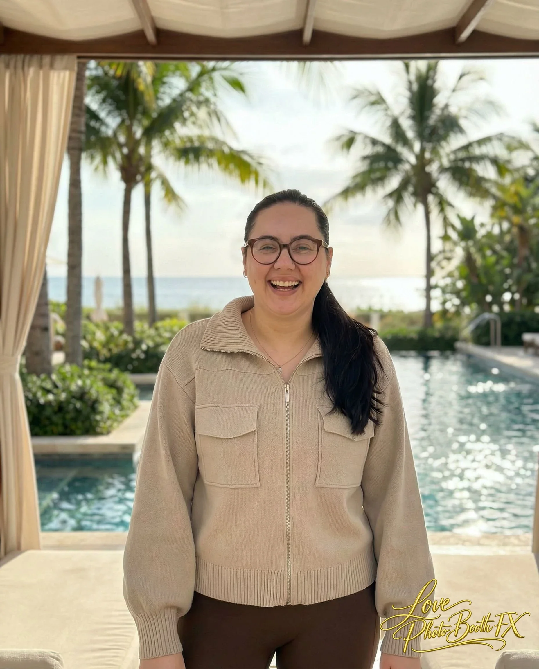 A woman smiling and laughing in a beige zip-up jacket standing outdoors near a swimming pool with palm trees and the ocean in the background.