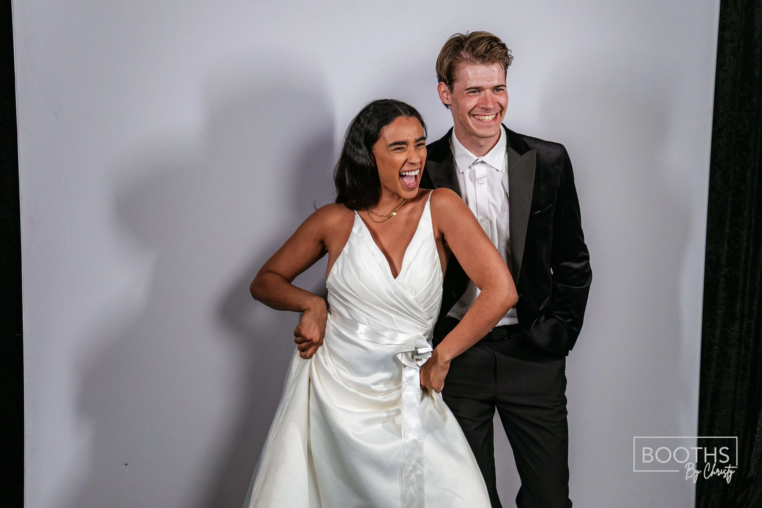A woman in a white dress and a man in a tuxedo are laughing together in front of a plain background.