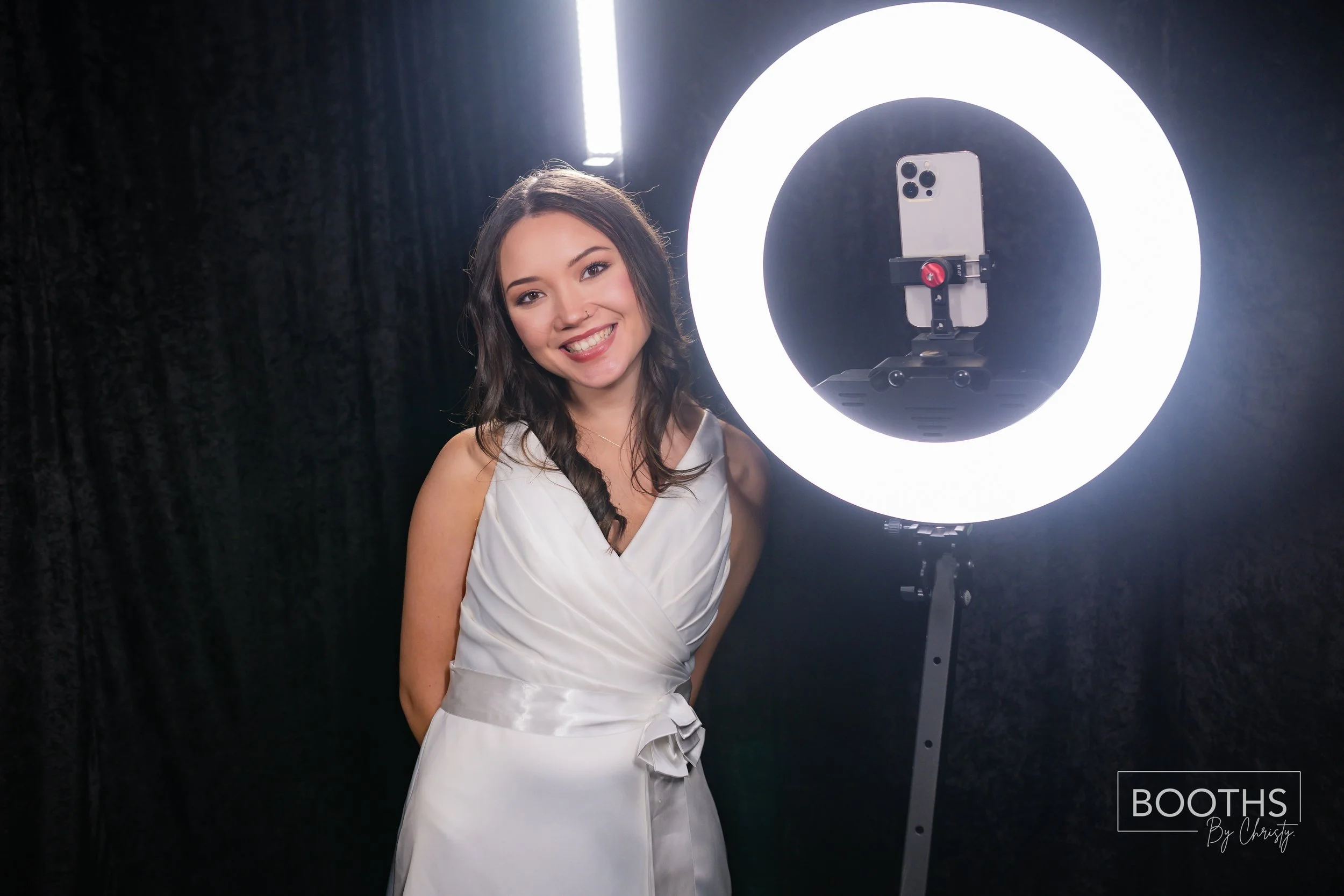 A woman in a white dress smiling near a ring light with a tablet mounted on a stand in a dark studio.