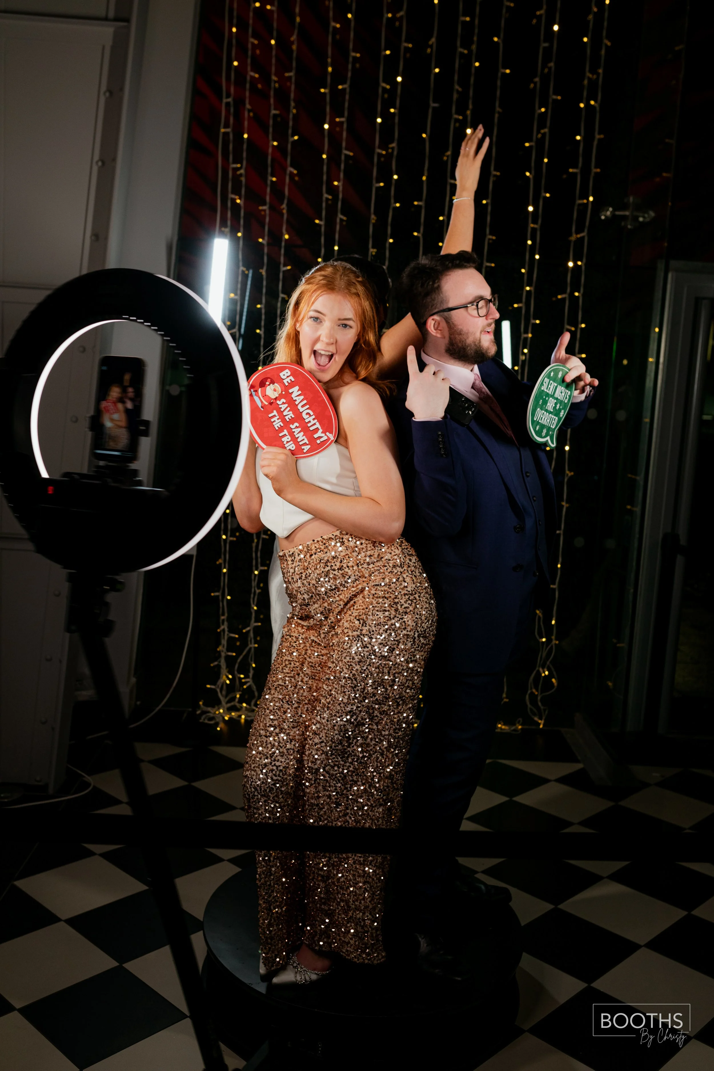 Two people standing in front of string lights taking a photo with a ring light, holding signs and posing playfully.