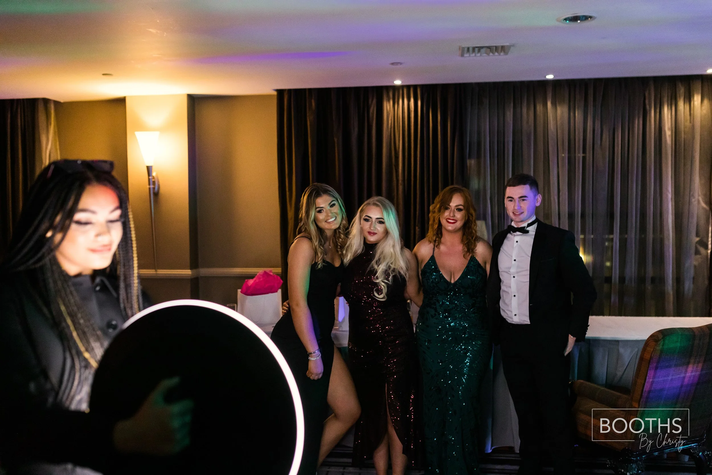Group of four people in formal attire posing for a photo at an indoor event, with a woman in the foreground taking a picture with a ring light.