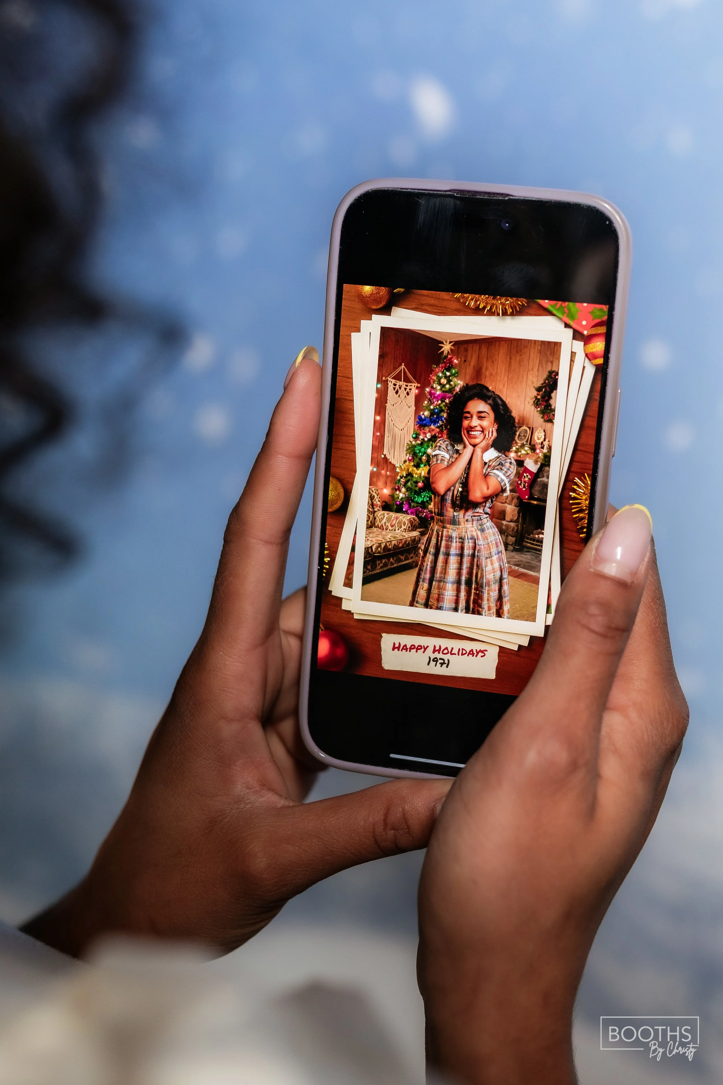 Person holding a smartphone displaying a Christmas photo of a woman smiling in front of a decorated Christmas tree. The photo is on a wooden table, surrounded by Christmas ornaments and decorations. The phone screen shows a label at the bottom saying