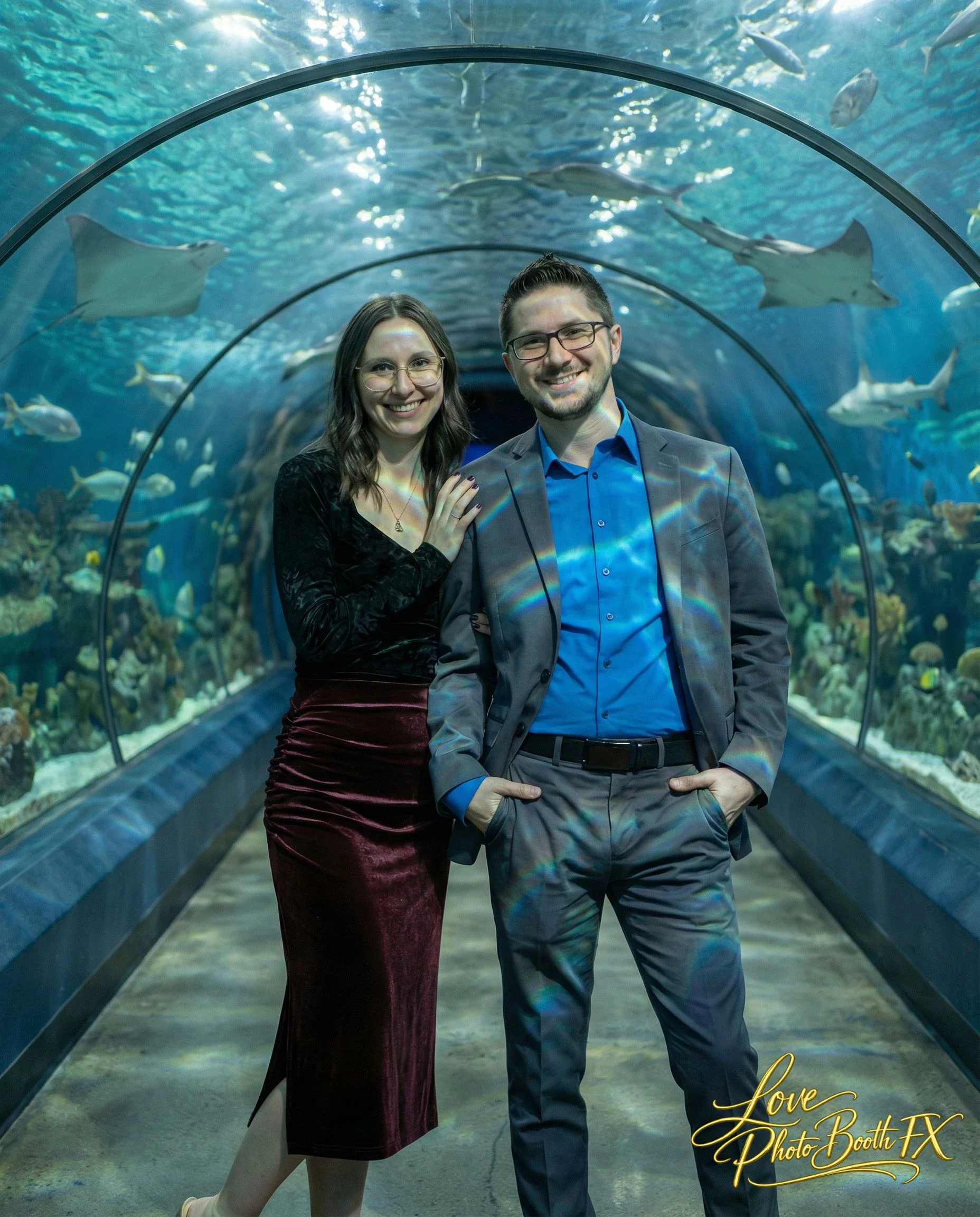 A smiling man and woman pose inside an aquarium tunnel with fish swimming overhead and around them.