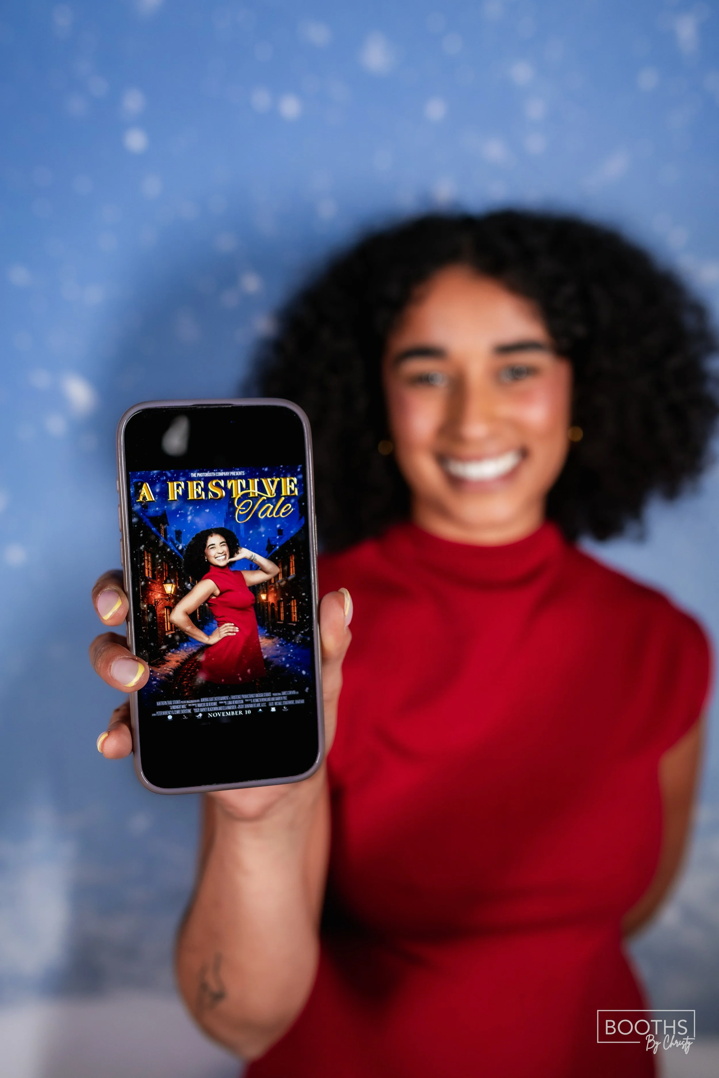 A smiling woman in a red dress holding a smartphone displaying a movie poster titled 'A Festive Tale' with a snowy street scene in the background.