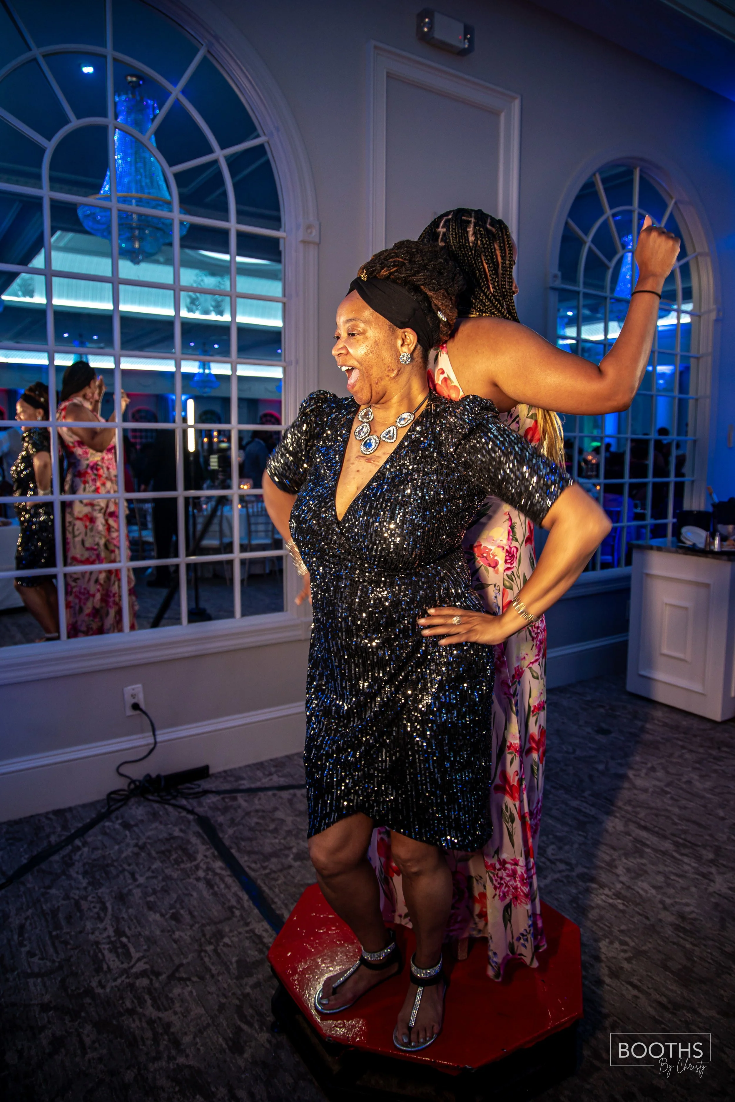 Two women dancing at a celebration. The woman in the foreground is wearing a black, sequined dress and silver jewelry, standing on a small dance platform inside a decorated venue with large arched windows and blue lighting.
