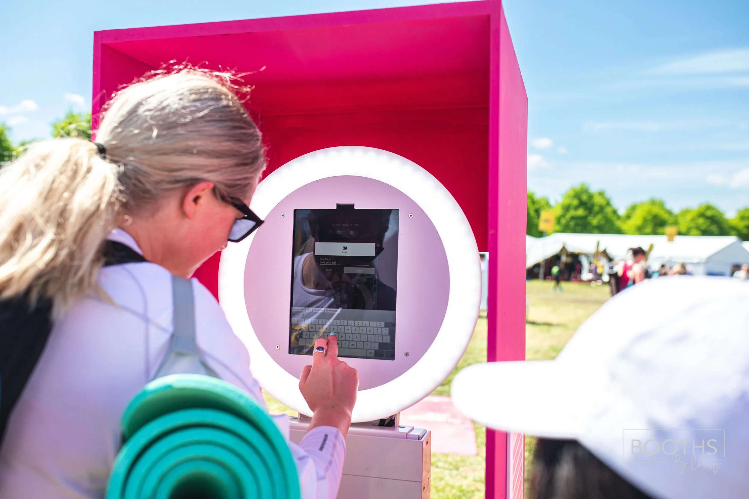 A person using a touchscreen kiosk outdoors with a pink frame and a ring light, at a festival or fair on a sunny day.