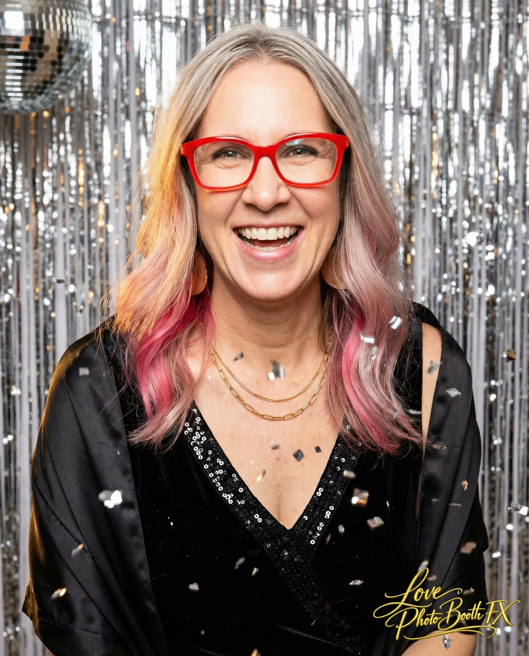 A smiling woman with blonde and pink hair, red glasses, and gold necklaces, standing in front of a metallic silver tinsel backdrop, decorated with confetti, during a celebration.