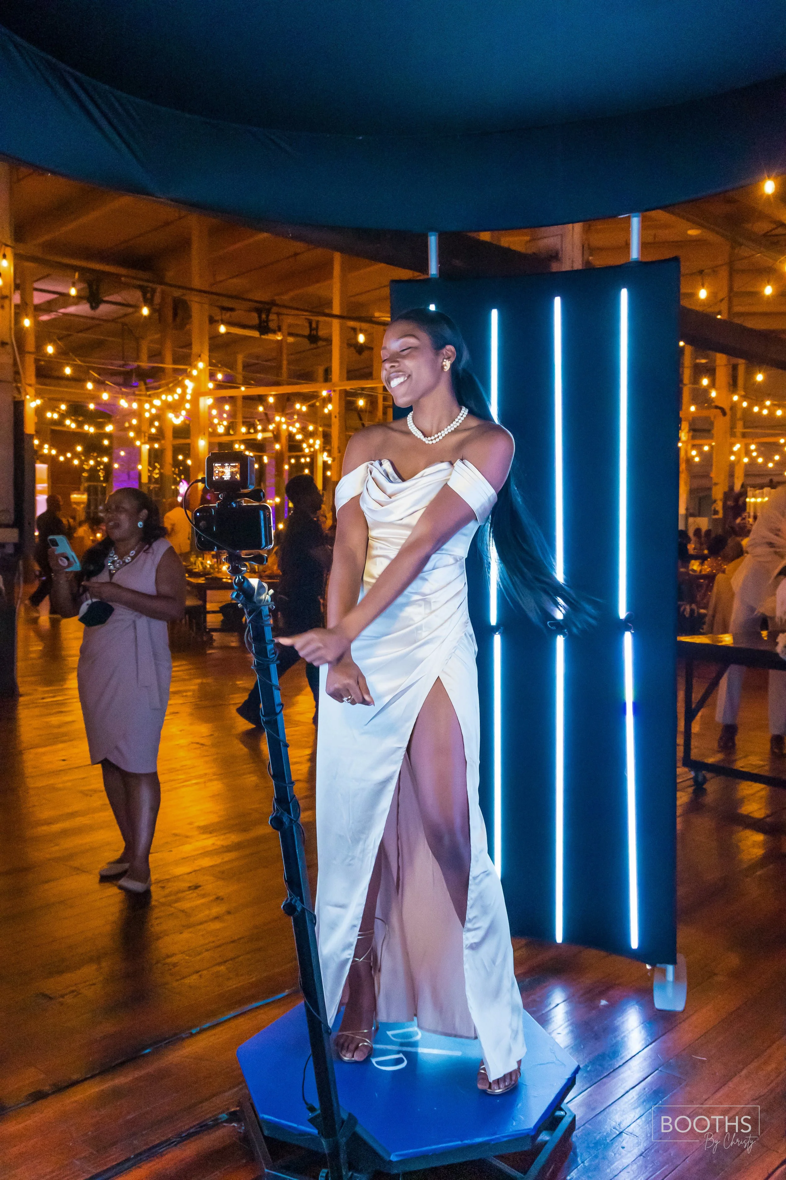 A woman in an elegant white gown with a thigh-high slit is standing on a platform, smiling and posing for photos at a formal event with string lights in the background.