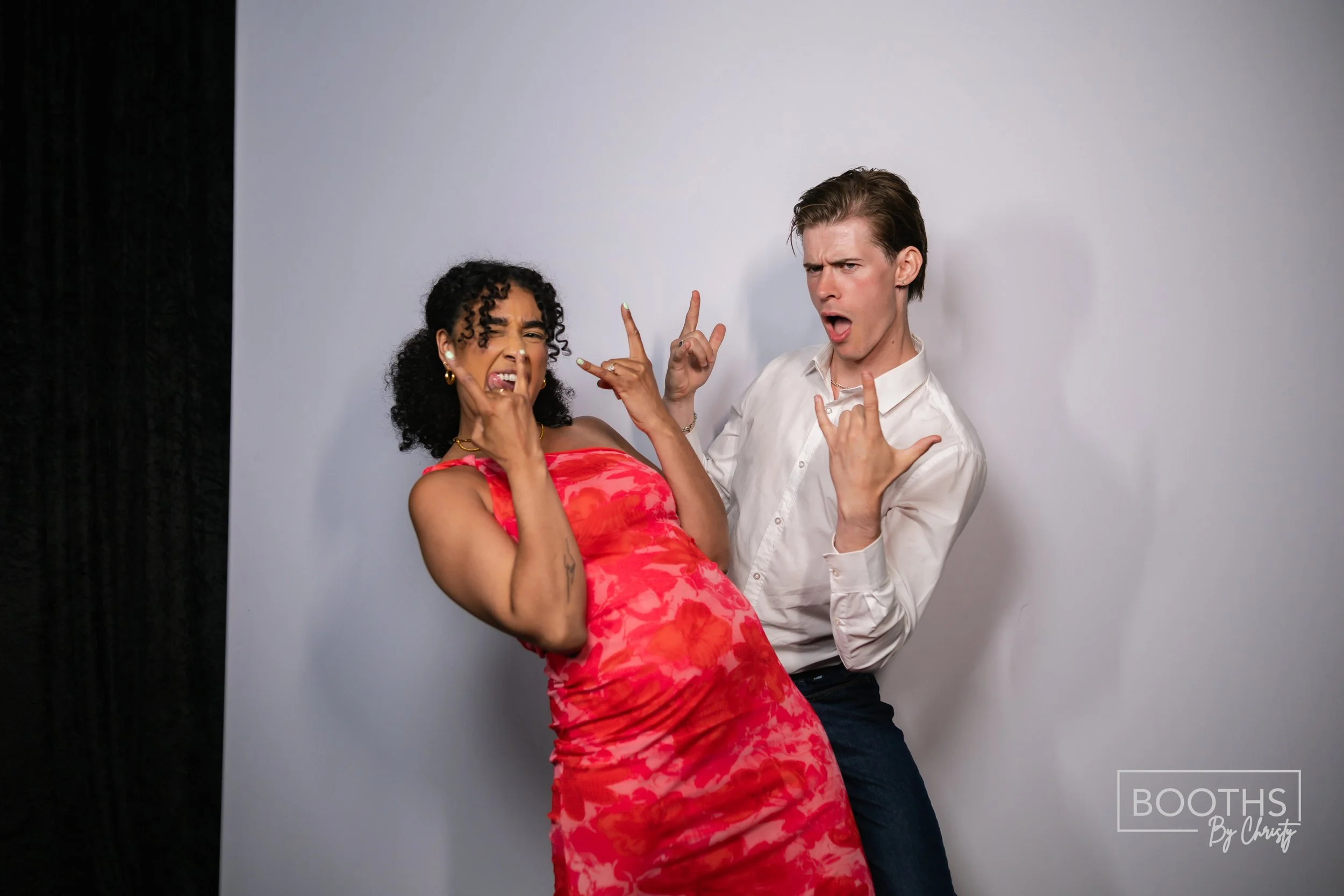 Two young adults making playful rock and roll gestures and facial expressions in a photo booth, with one woman wearing a red dress and a man in a white shirt, standing in front of a plain backdrop.