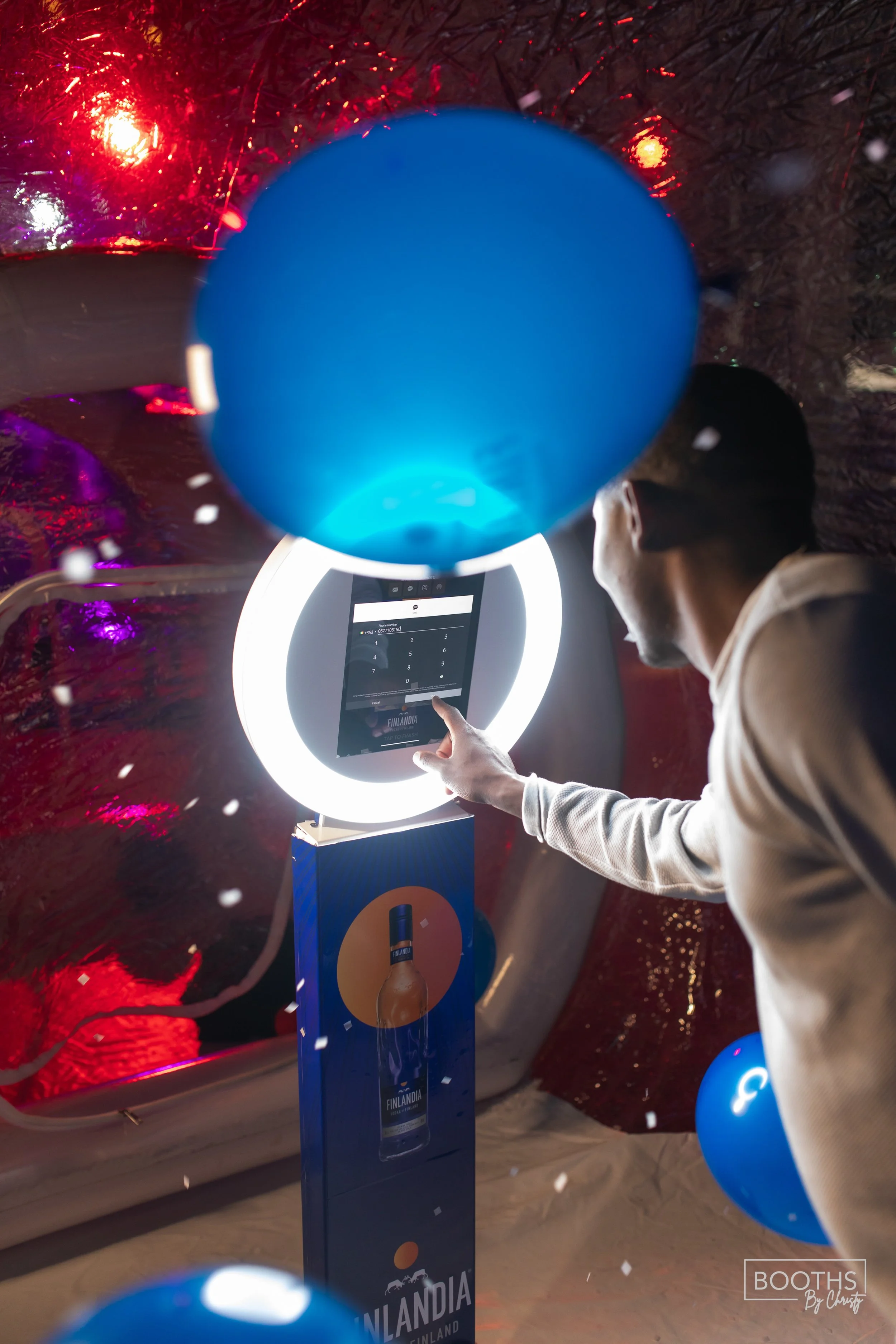 Person using a digital kiosk with a bright circular ring light at an indoor event, surrounded by blue balloons and confetti, with a promotional display for Finlandia vodka in the foreground.