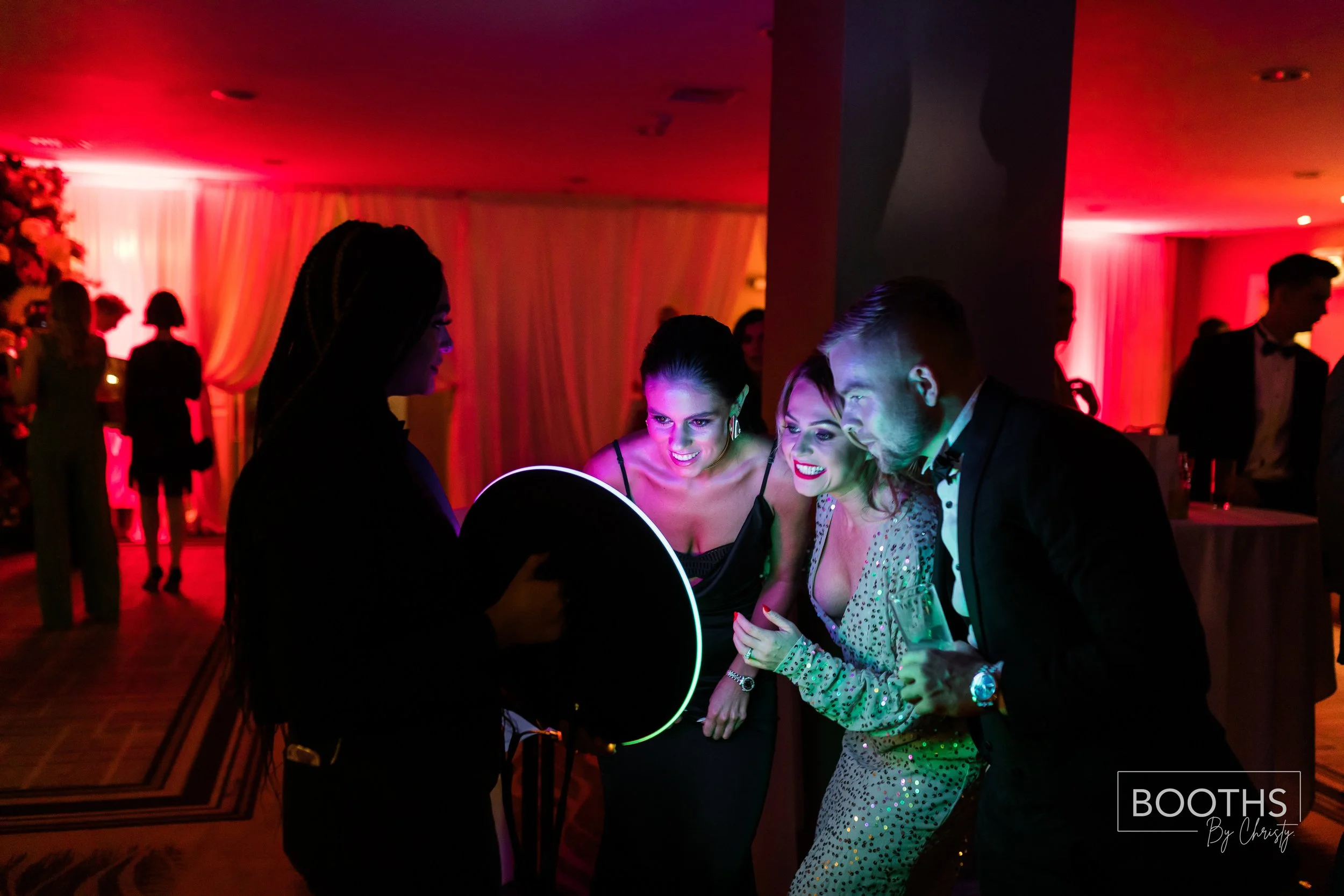 A group of four people at a party, smiling and looking at a tablet, with colorful lighting in the background.
