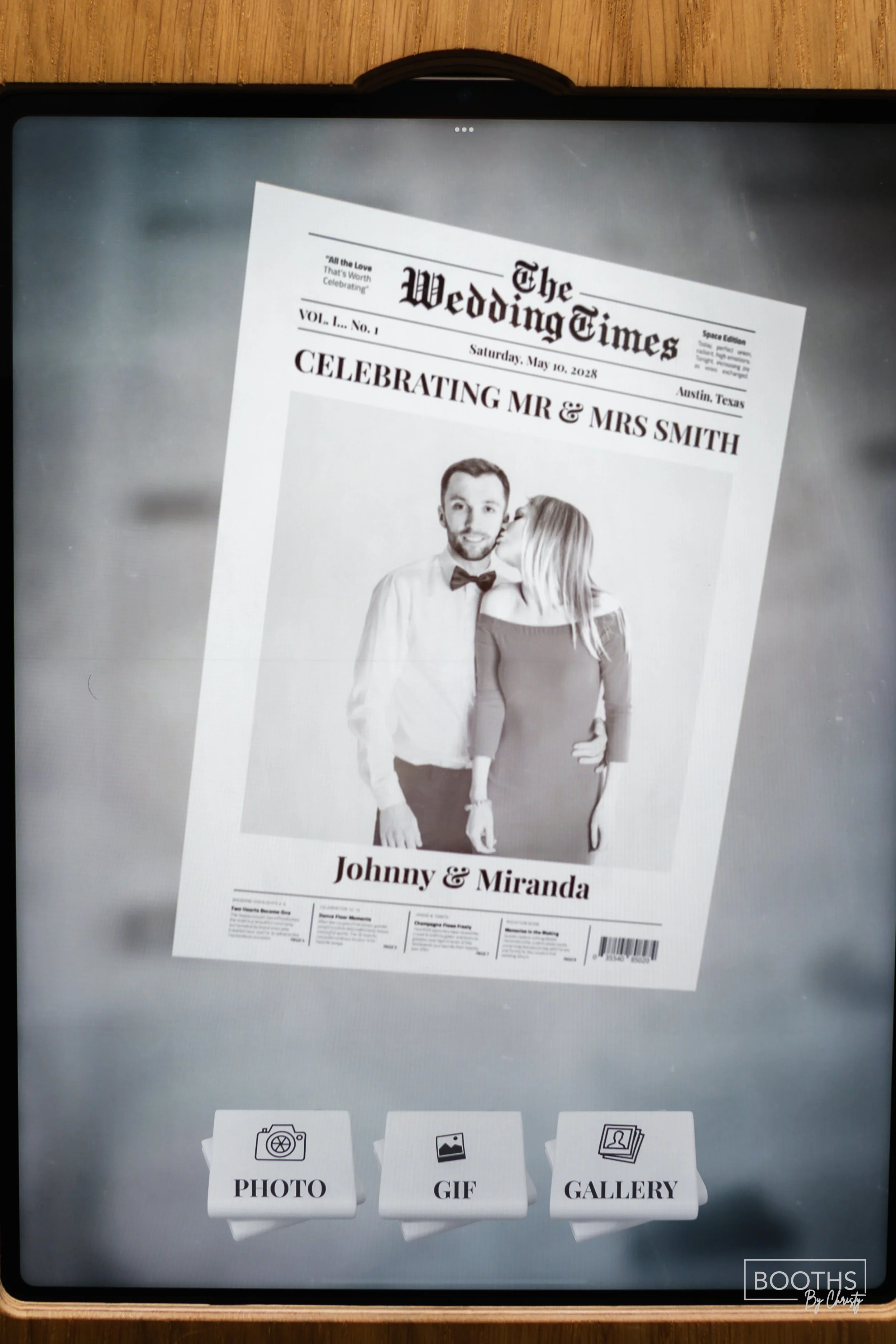 A digital screen displaying a photo booth setup with a black and white wedding newspaper poster showing a couple, Johnny and Miranda, on the front cover. The poster has the date May 10, 2028, and the location Austin Texas. There are three options at