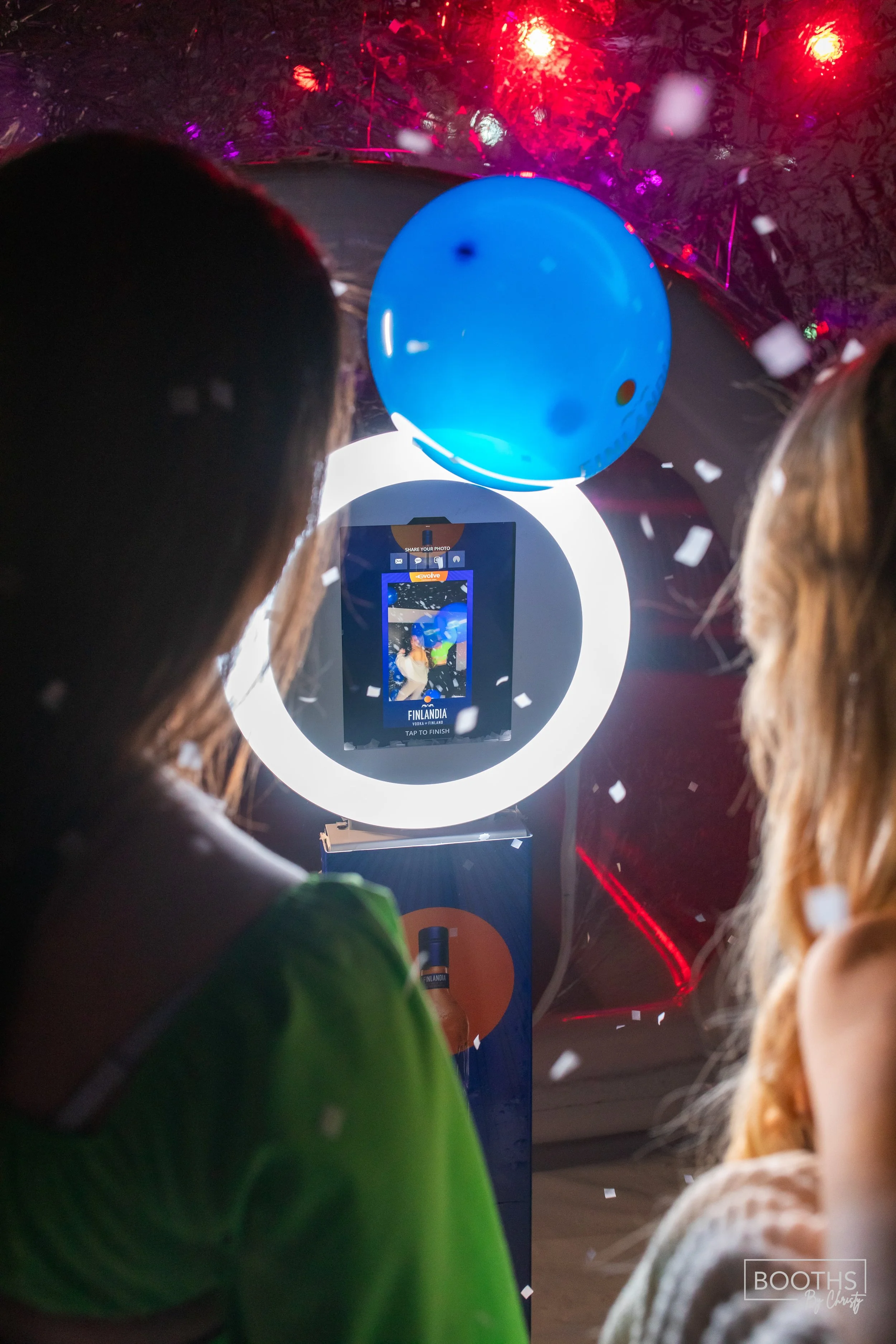 Two women at a photo booth, with a blue balloon above them and confetti falling, as they take a picture on a digital camera with a bright ring light in the background.