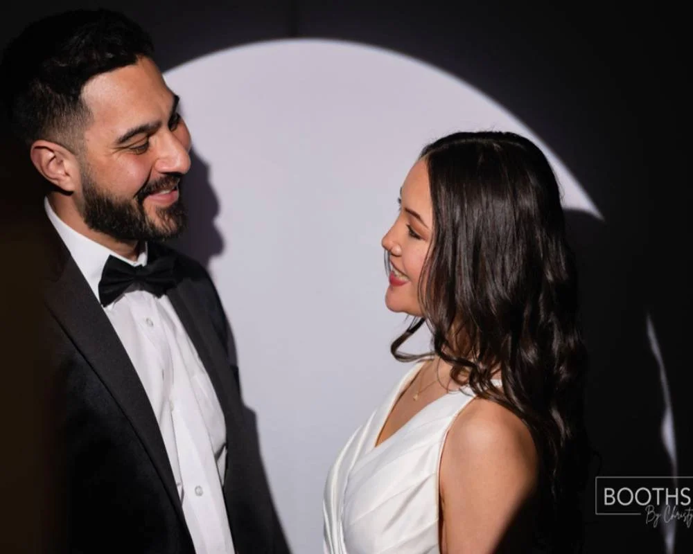 A man in a tuxedo and a woman in a white dress smiling at each other in a photo with a black and white background.