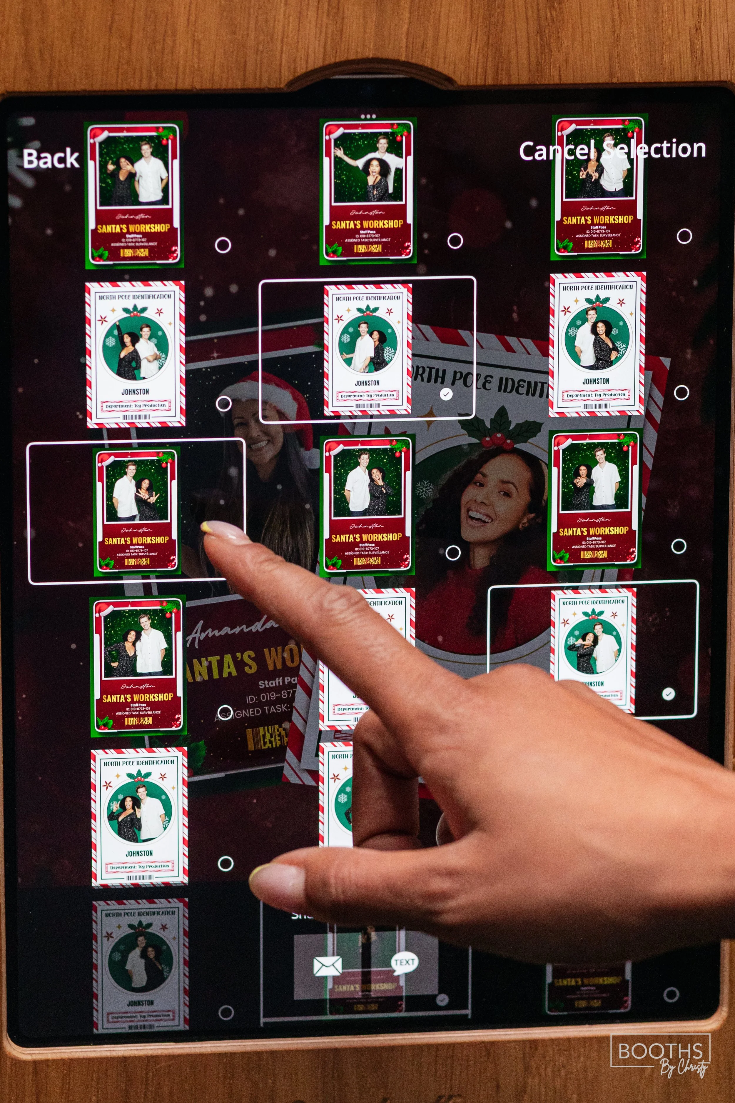A person's hand touching a photo booth screen displaying holiday-themed photo IDs for a Christmas Santa's workshop event, with various ID cards arranged on the screen.
