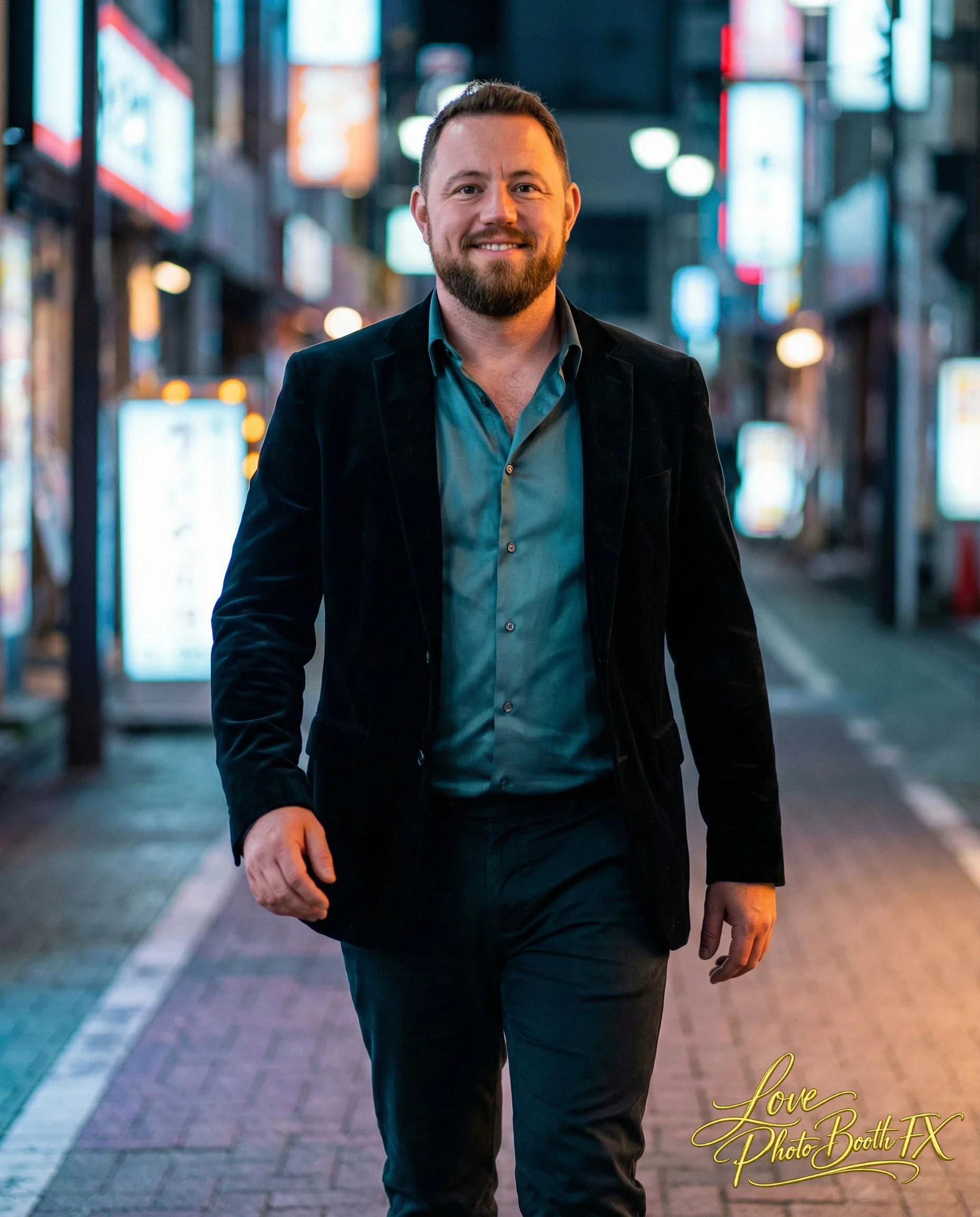 A man with a beard and short hair walking in an urban area at night, dressed in a black blazer and teal shirt, smiling at the camera, with city lights and signs in the background.