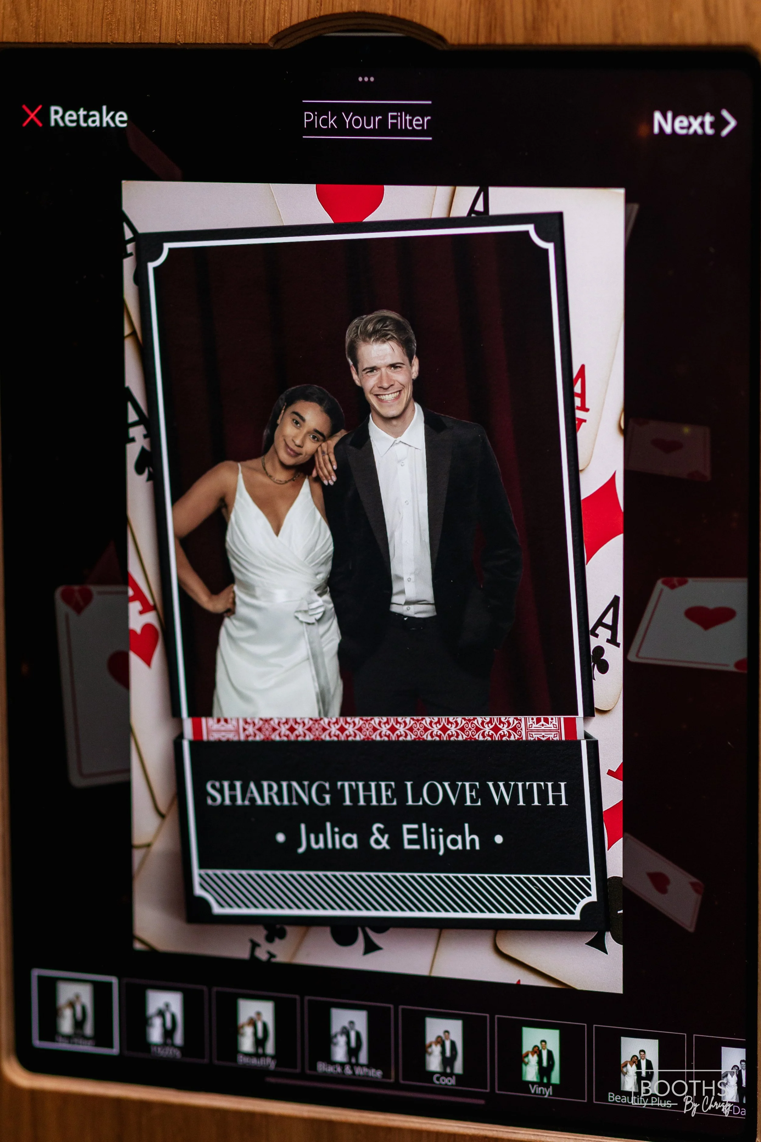 A photo booth screen displaying a picture of a woman in a white dress and a man in a black tuxedo, with a shadowed red curtain background. The overlay reads "Sharing the Love with Julia & Elijah".