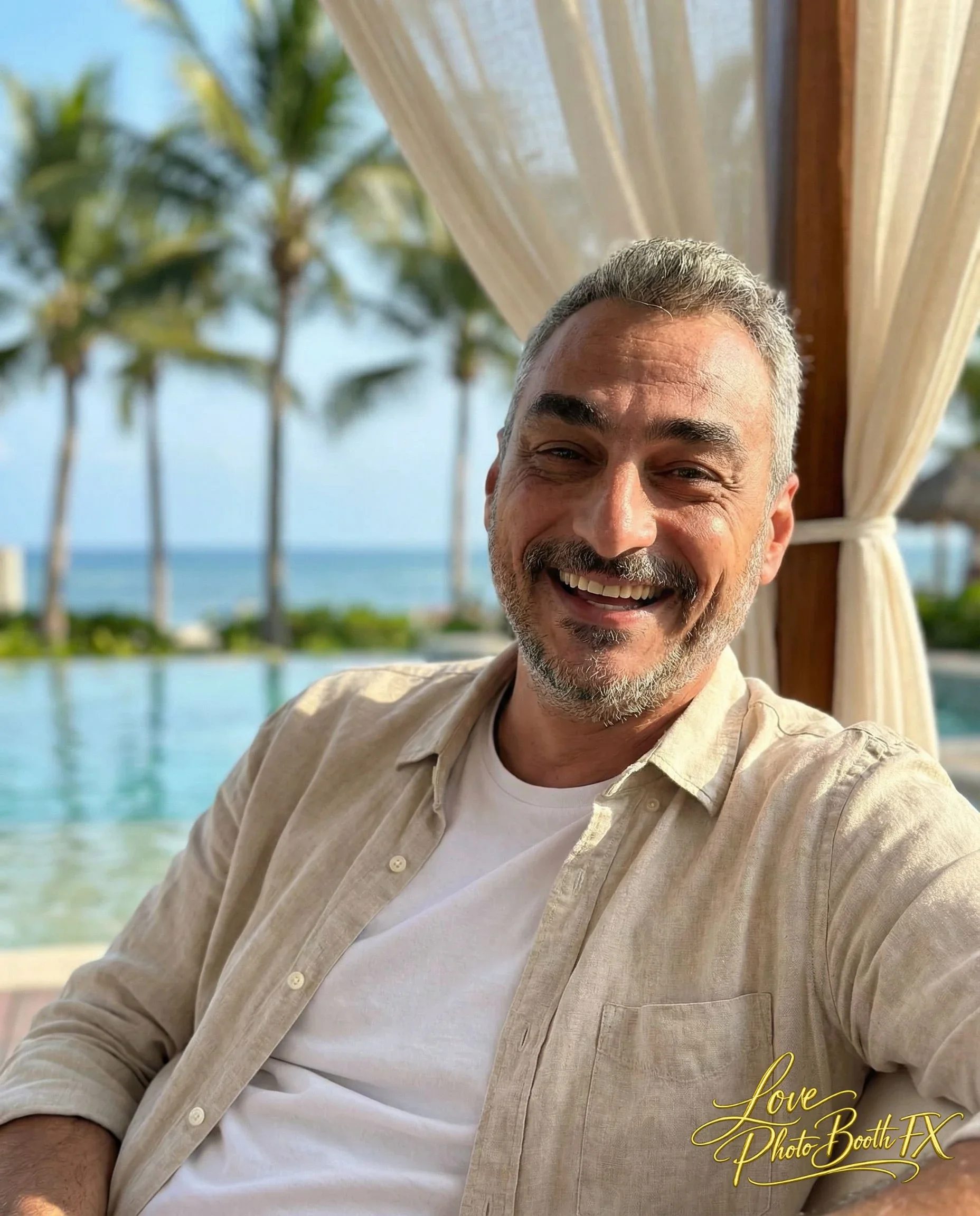 A smiling man with gray hair and a beard taking a selfie outdoors near a pool, with palm trees and the ocean in the background, under a curtain-draped cabana.