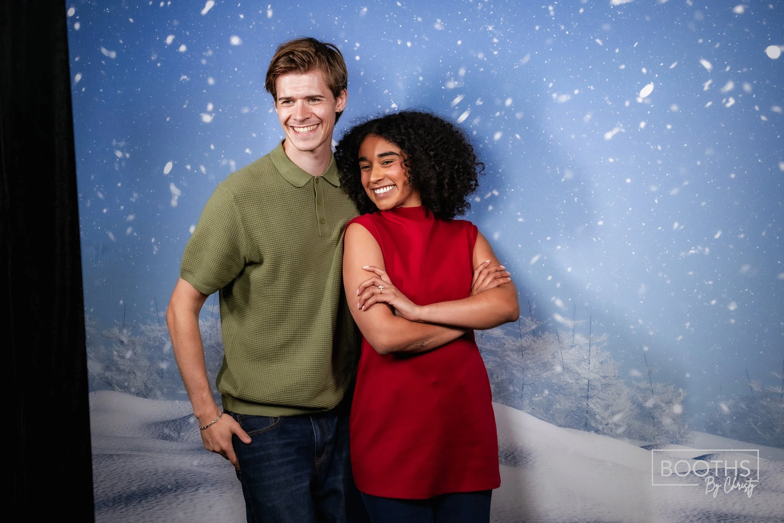 Smiling couple standing together in front of a snowy winter-themed backdrop, with the man wearing a green polo shirt and the woman wearing a red sleeveless top.