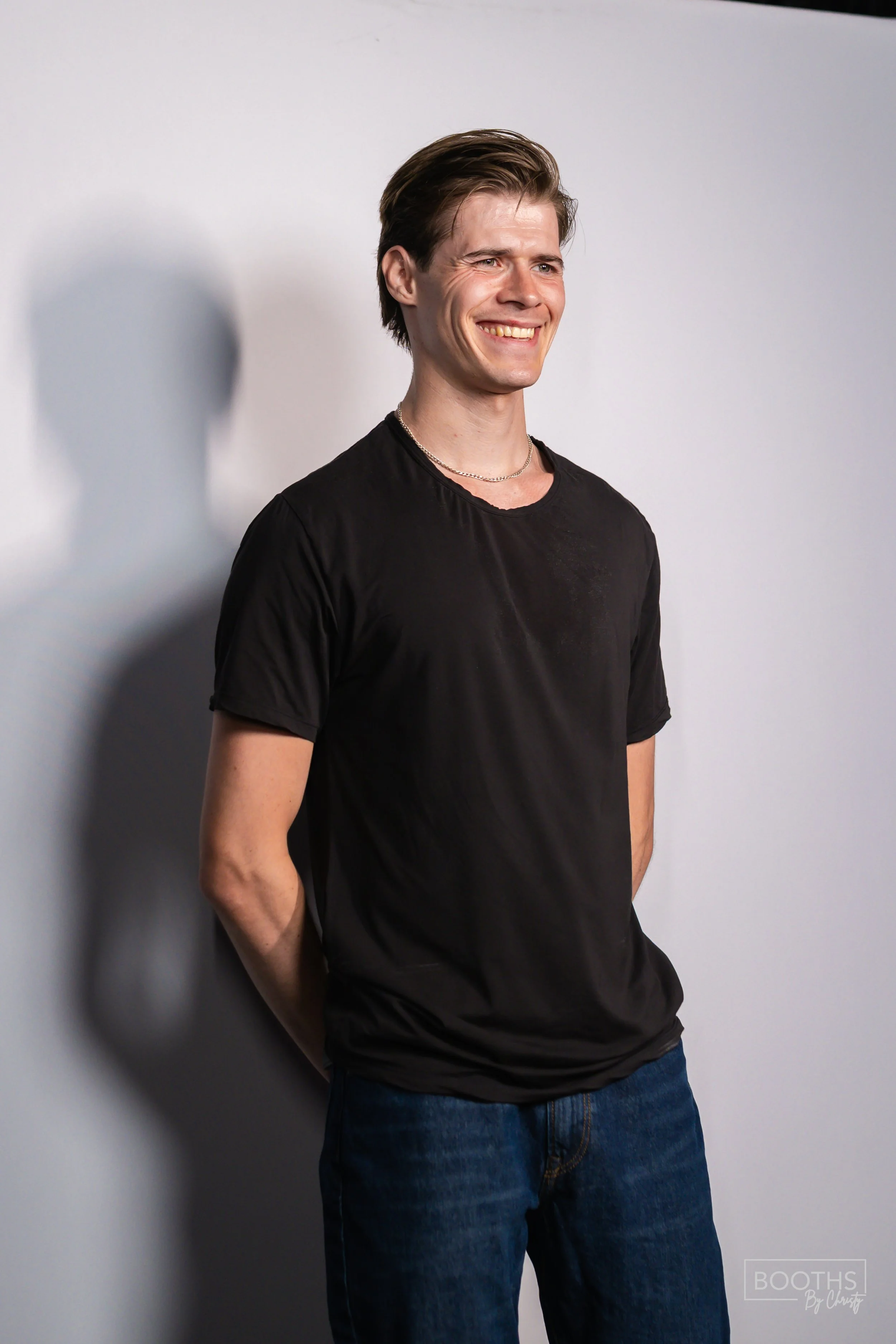 A young man with brown hair, wearing a black t-shirt and blue jeans, standing against a white background, smiling and looking to the side.