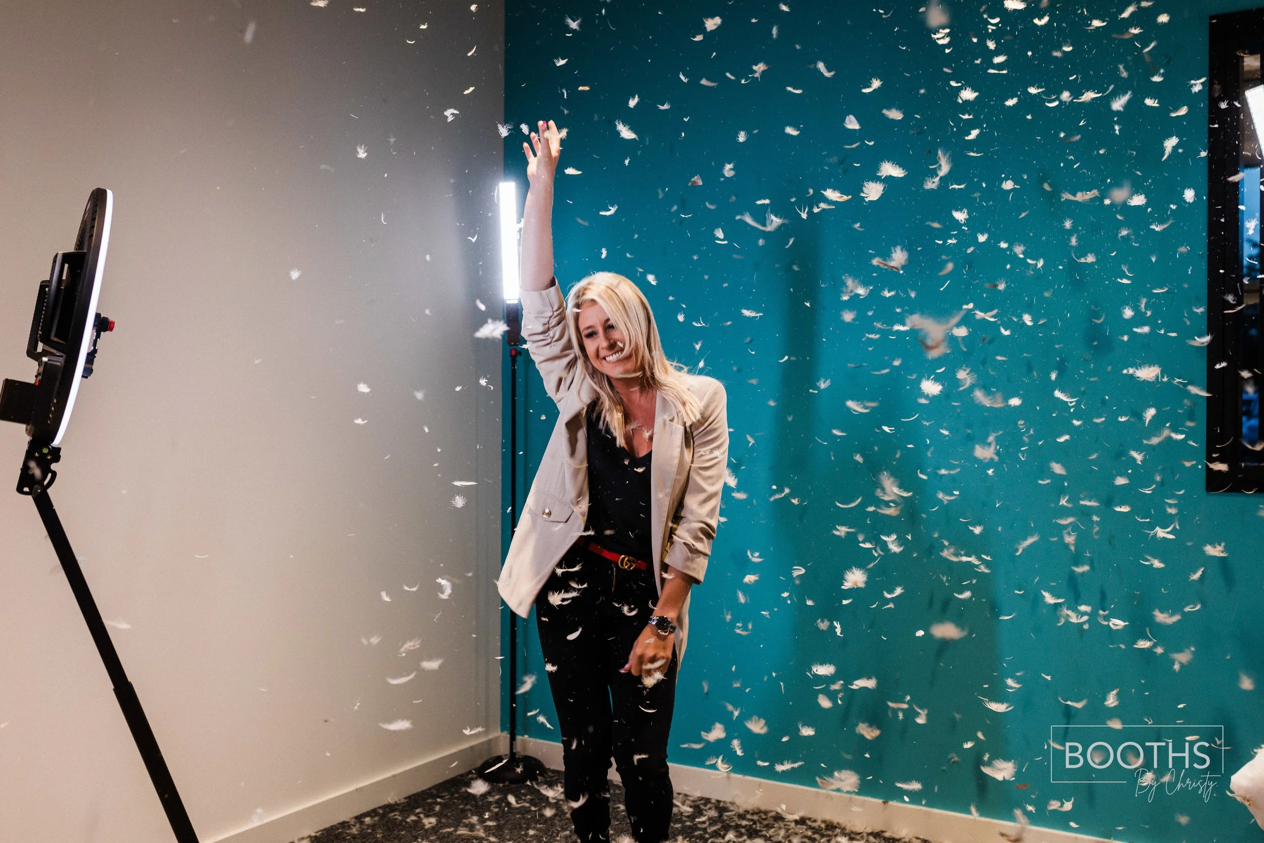 A woman in a beige blazer and black outfit smiling and throwing feathers in the air at a photo booth with a blue and gray wall background.