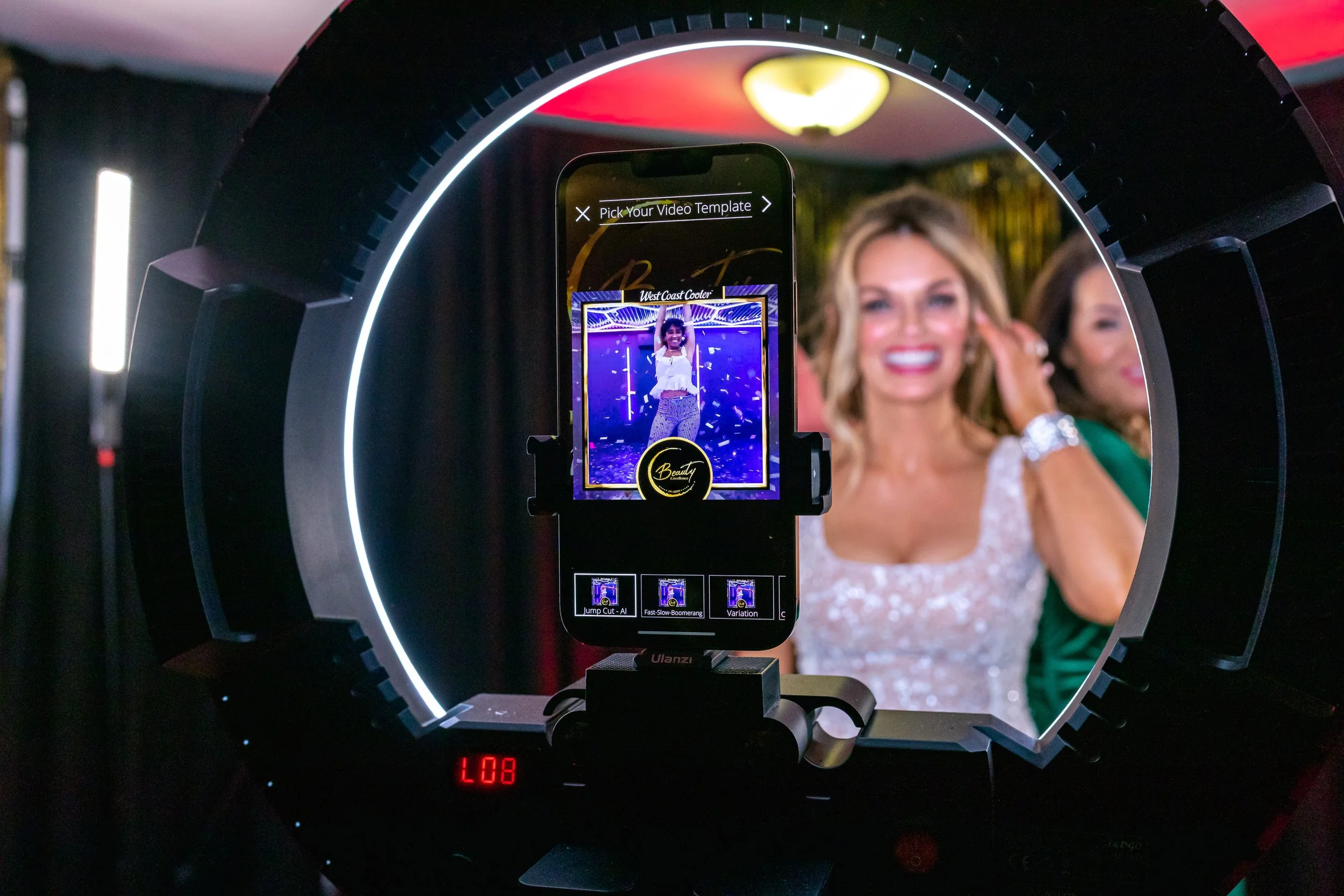 A photo booth setup capturing a smiling woman in a white lace dress with a woman in green behind her, with a smartphone on a ring light displaying the scene.