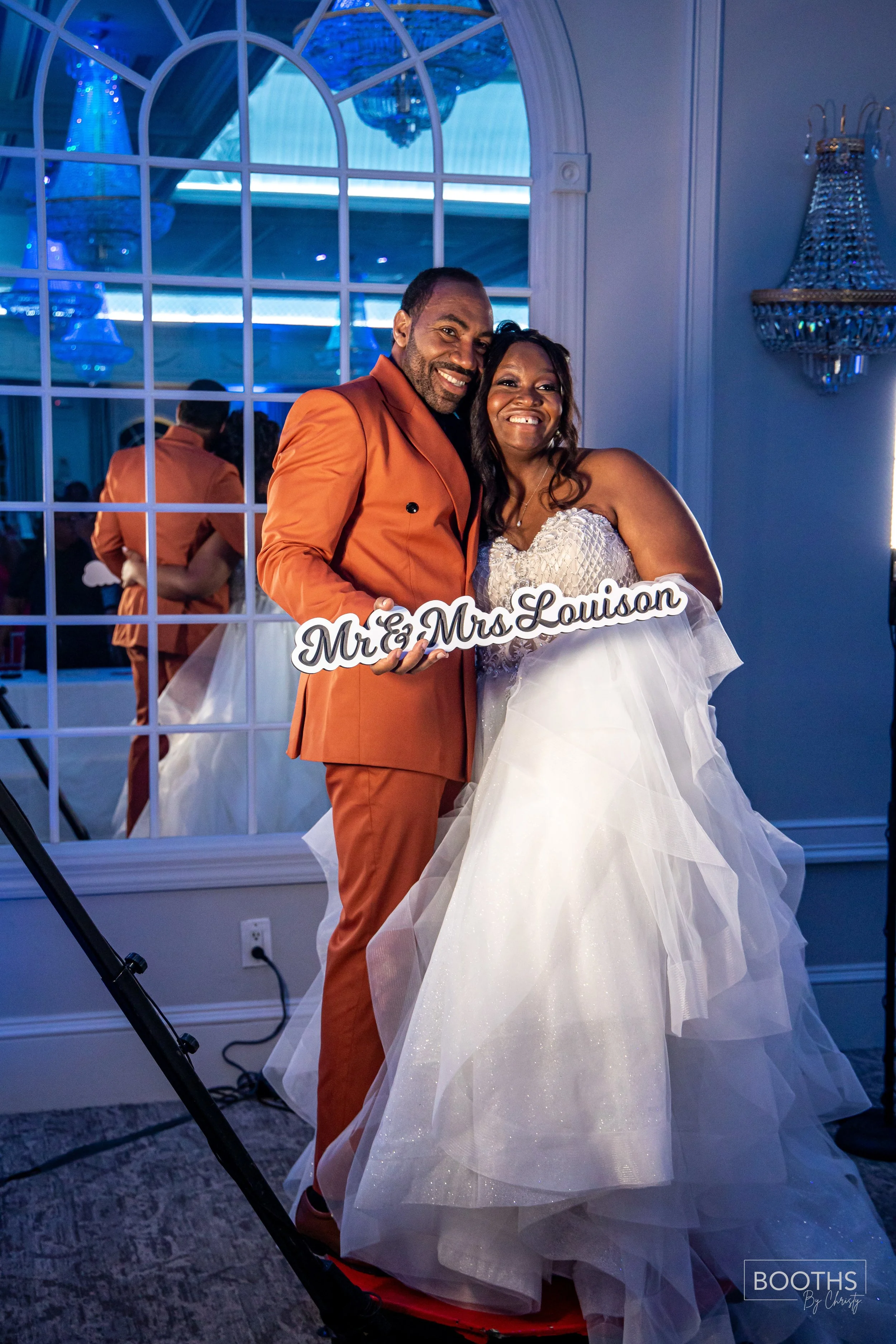 A newlywed couple smiling at a wedding reception, holding a sign that reads 'Mr. & Mrs. Louison,' with a large mirror and chandelier in the background.