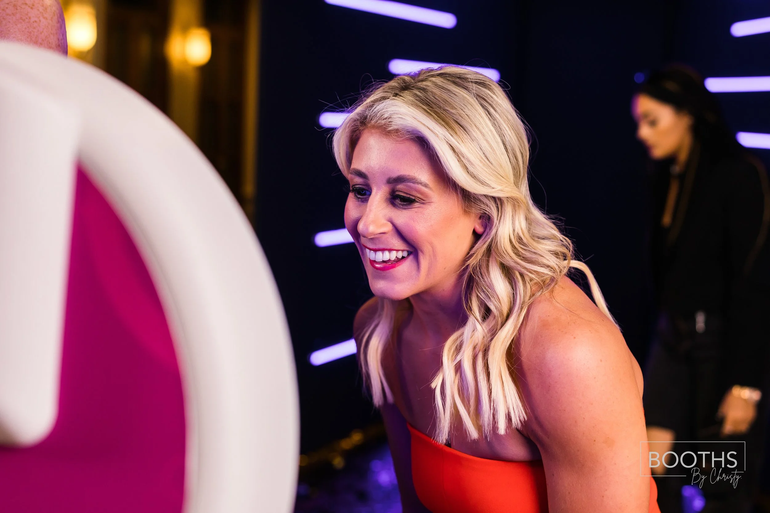 A woman with blonde hair and a red dress smiling at a photo booth at an event with neon lighting.