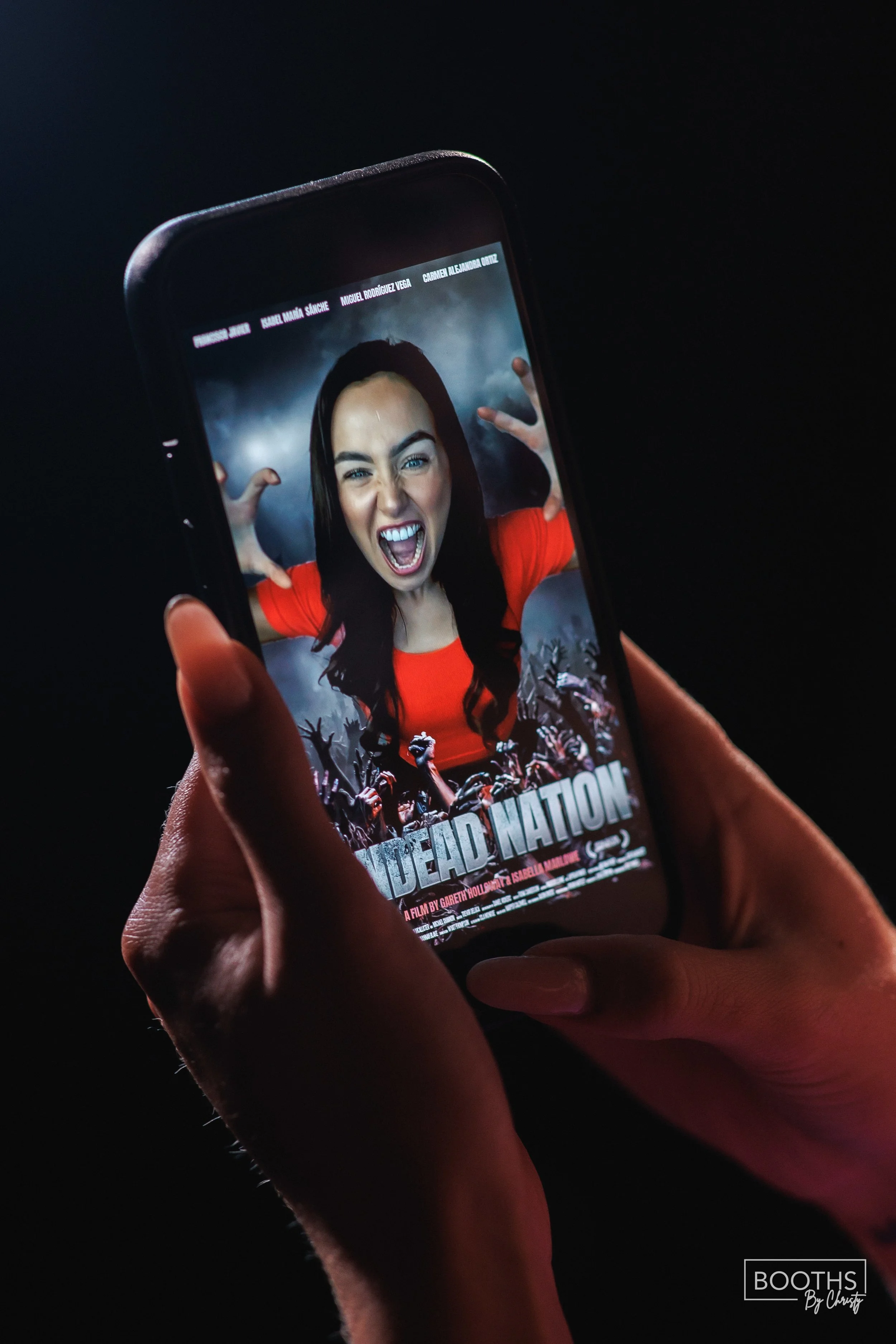 A person holding a smartphone displaying the movie poster for 'Dead Nation,' showing a woman with dark hair, wearing a red shirt, making a crazy face with her hands near her head, surrounded by zombies.