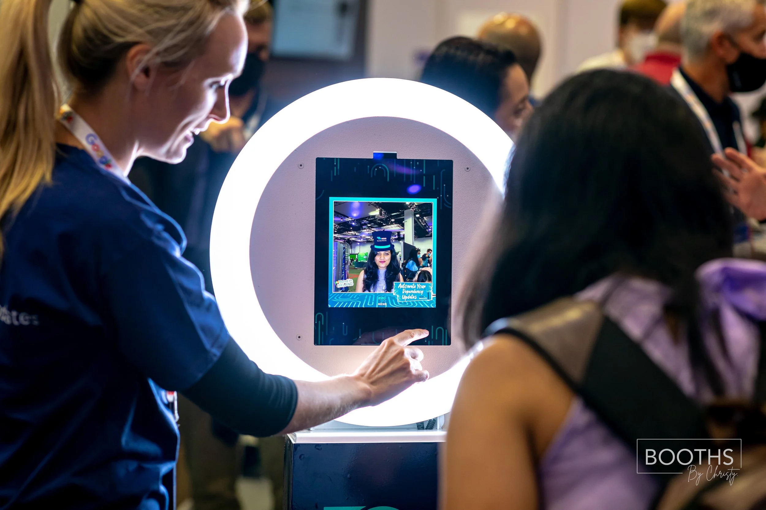 A woman is using a photo booth with a circular light, capturing her image on a digital screen, at a technology or event expo.