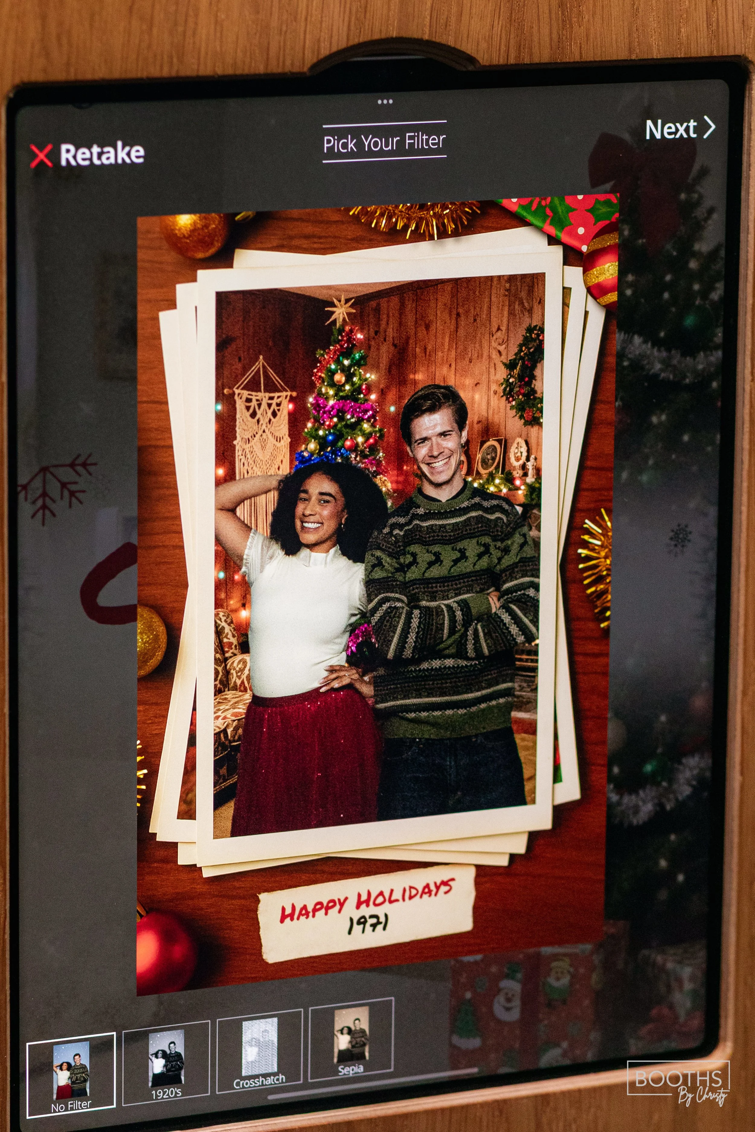 A photo of a smiling woman and man standing in front of a decorated Christmas tree, with a wooden wall and holiday decorations in the background. The photo is displayed on a digital screen with Christmas ornaments and a note that reads "HAPPY HOLIDAY