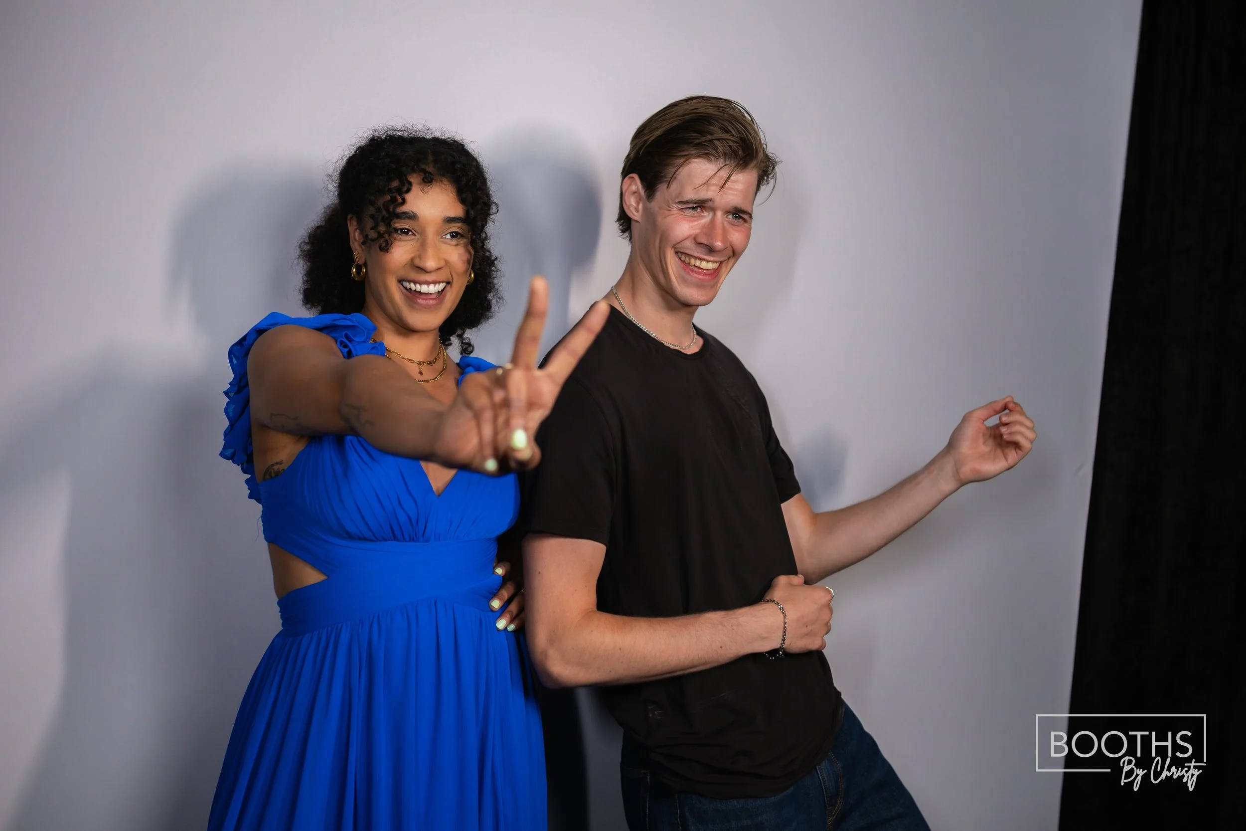 Two people smiling and posing for a photo. The woman on the left, wearing a blue dress, makes a peace sign with her hand. The man on the right, dressed in a black t-shirt, also smiles, standing in front of a gray background.