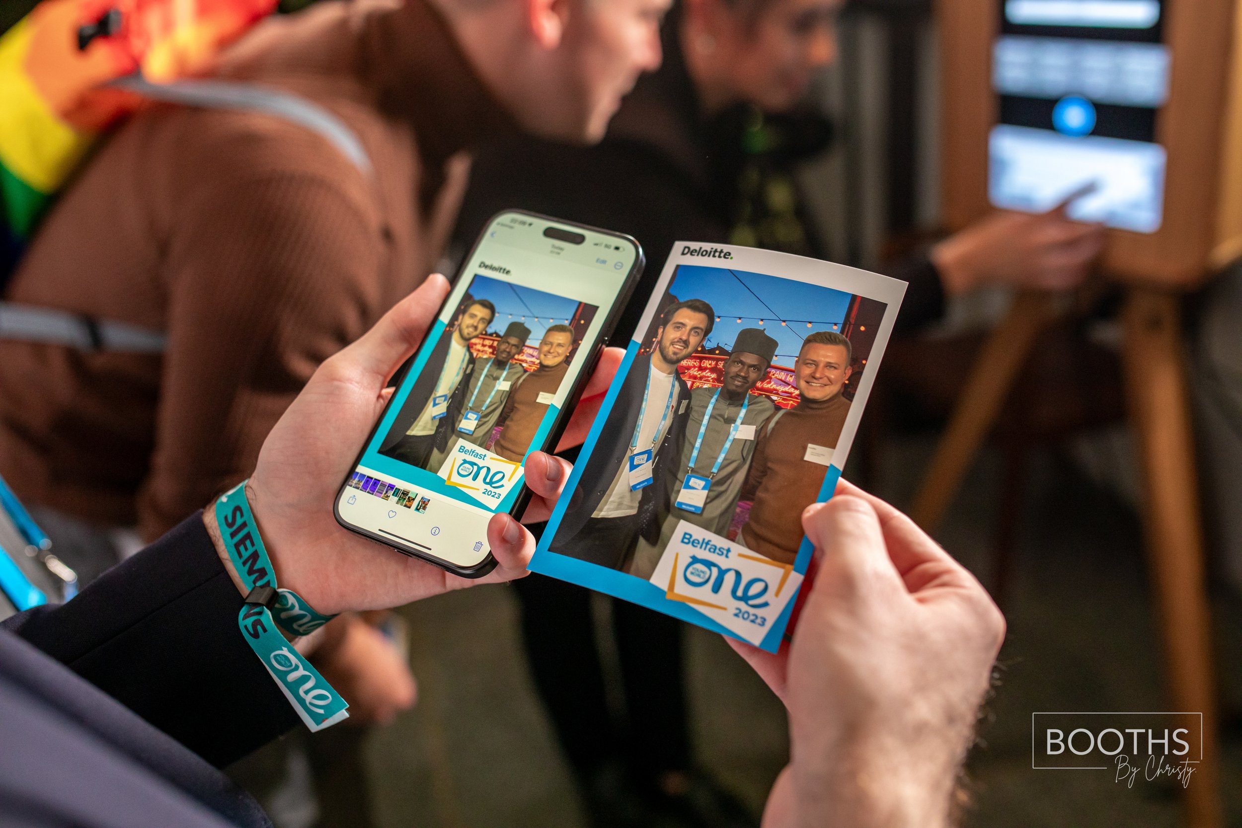 Person taking a photo of a printed photo using a smartphone at a conference. The printed photo shows three men posing outdoors, with a