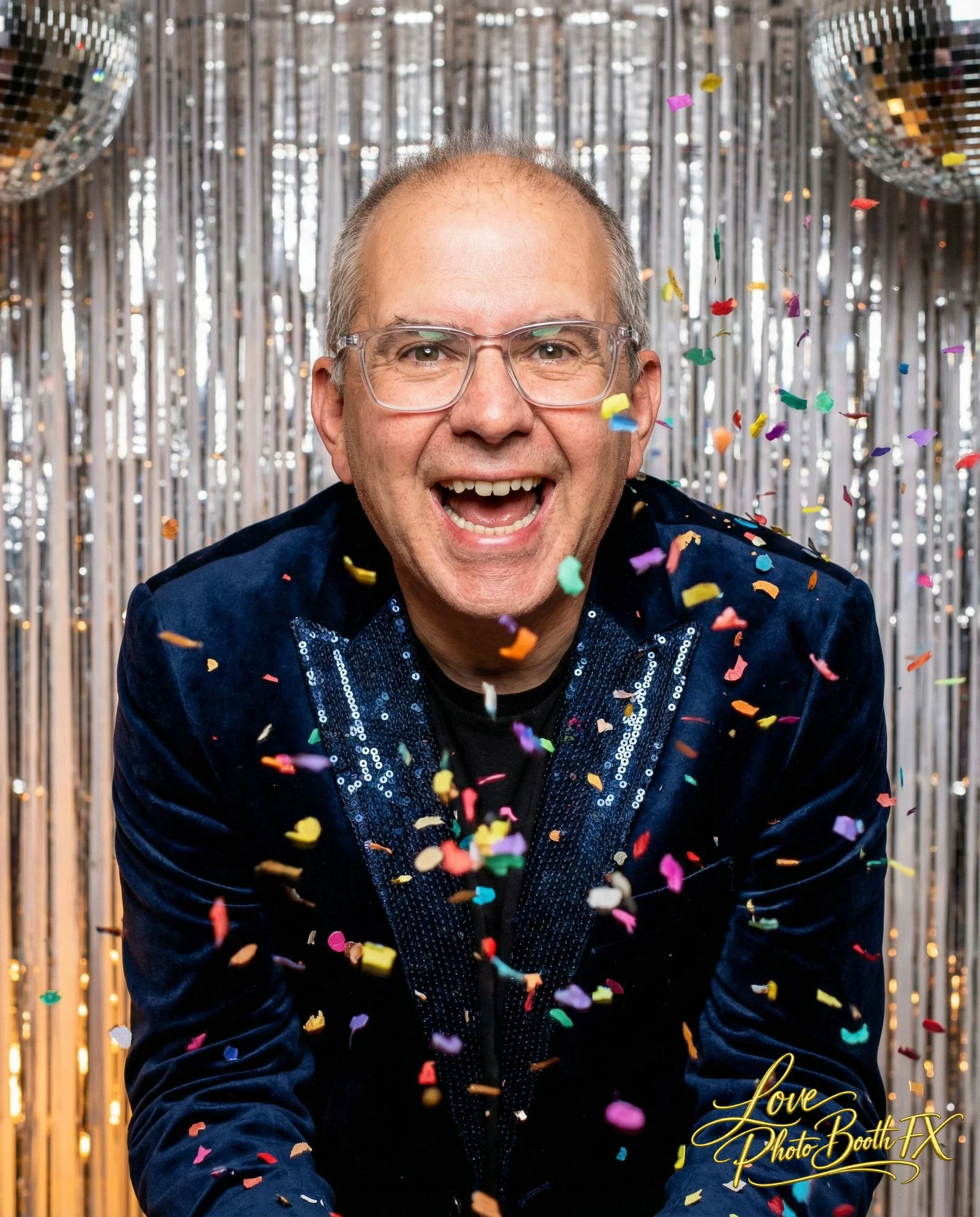 Man smiling with glasses, wearing a dark blue velvet blazer with sequins, surrounded by falling colorful confetti and silver metallic streamers, with a silver tinsel backdrop and two disco balls.