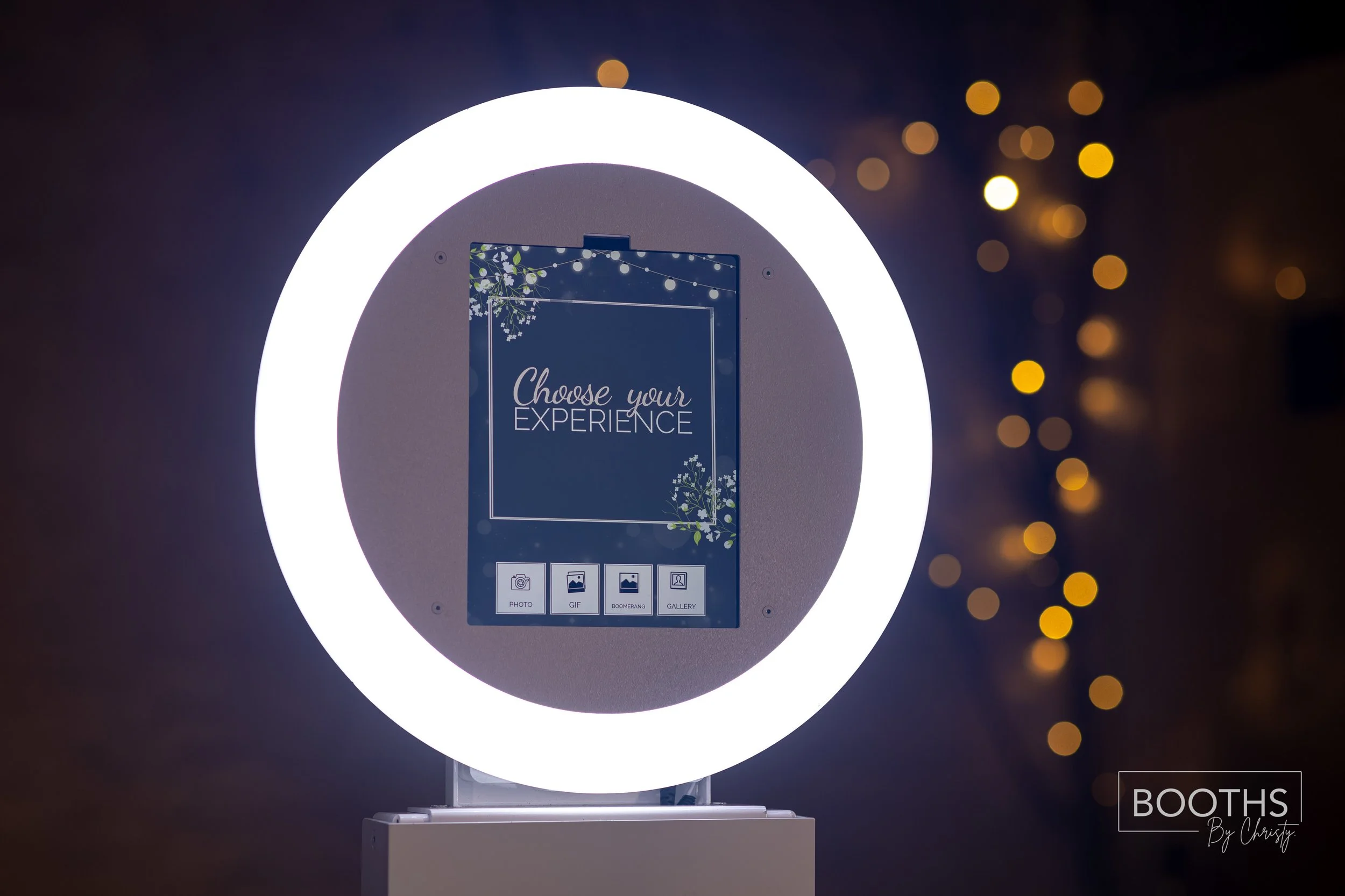 A photo booth with a circular ring light illuminating a digital screen that displays the message “Choose your EXPERIENCE” with options for photo, gif, boomerang, and gallery, against a dark background with bokeh lights.