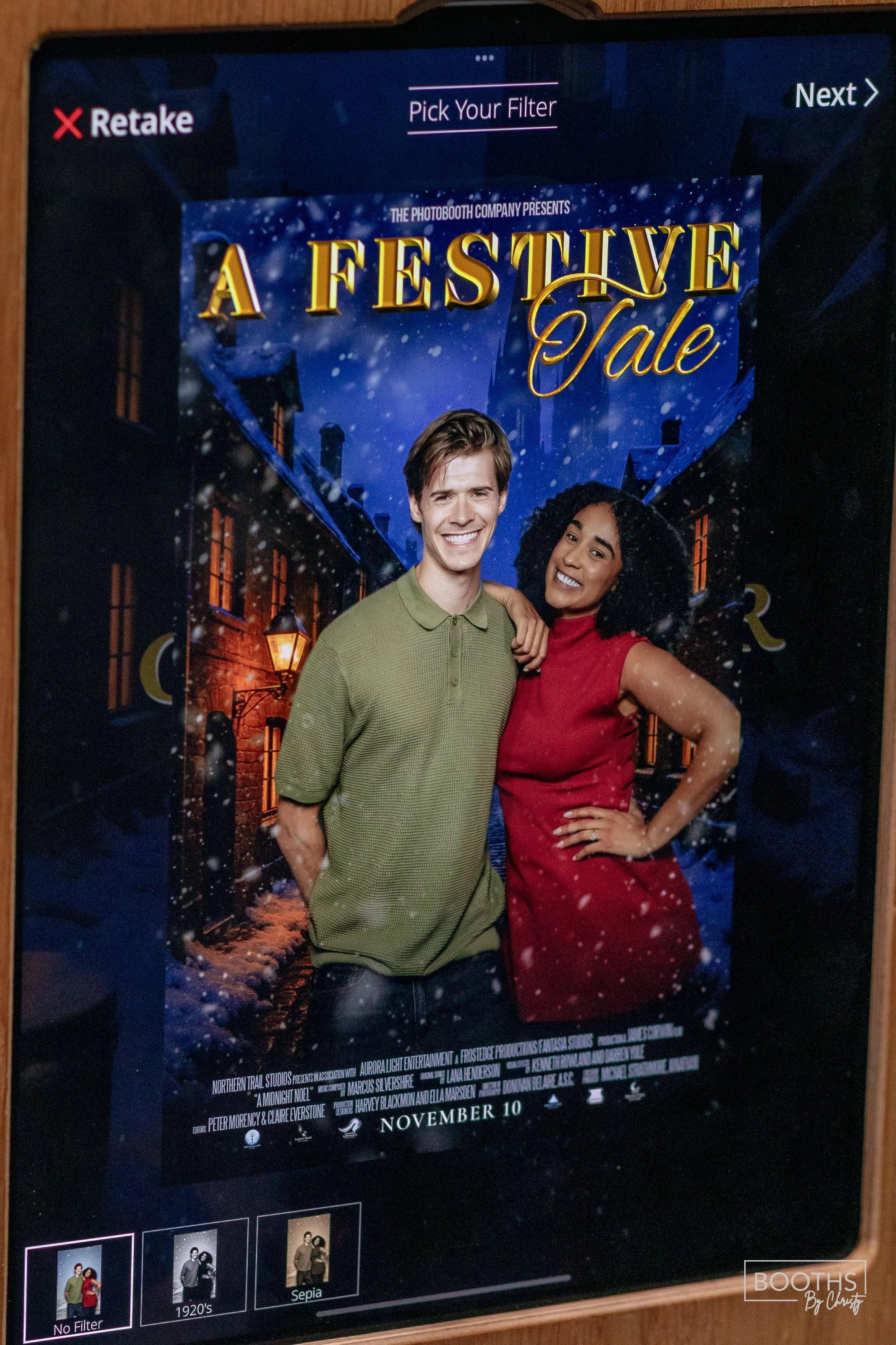 A digital screen displaying a photo booth poster for a holiday-themed event called "A Festive Tale," featuring a smiling man and woman standing together against a snowy street background with houses and street lamps. The poster indicates the event is