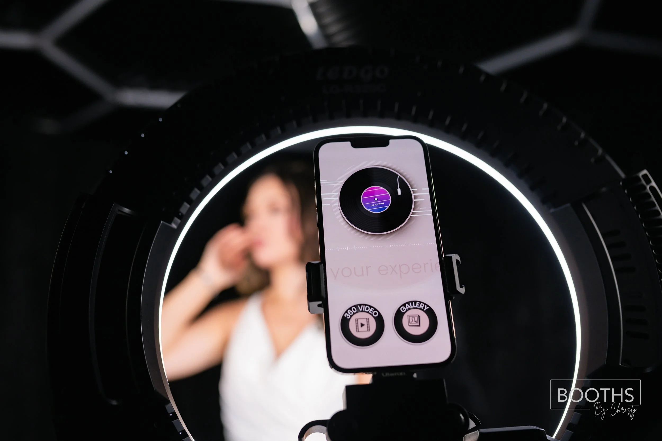 A woman is posing for a photo in front of a ring light with a phone on a tripod capturing the image.