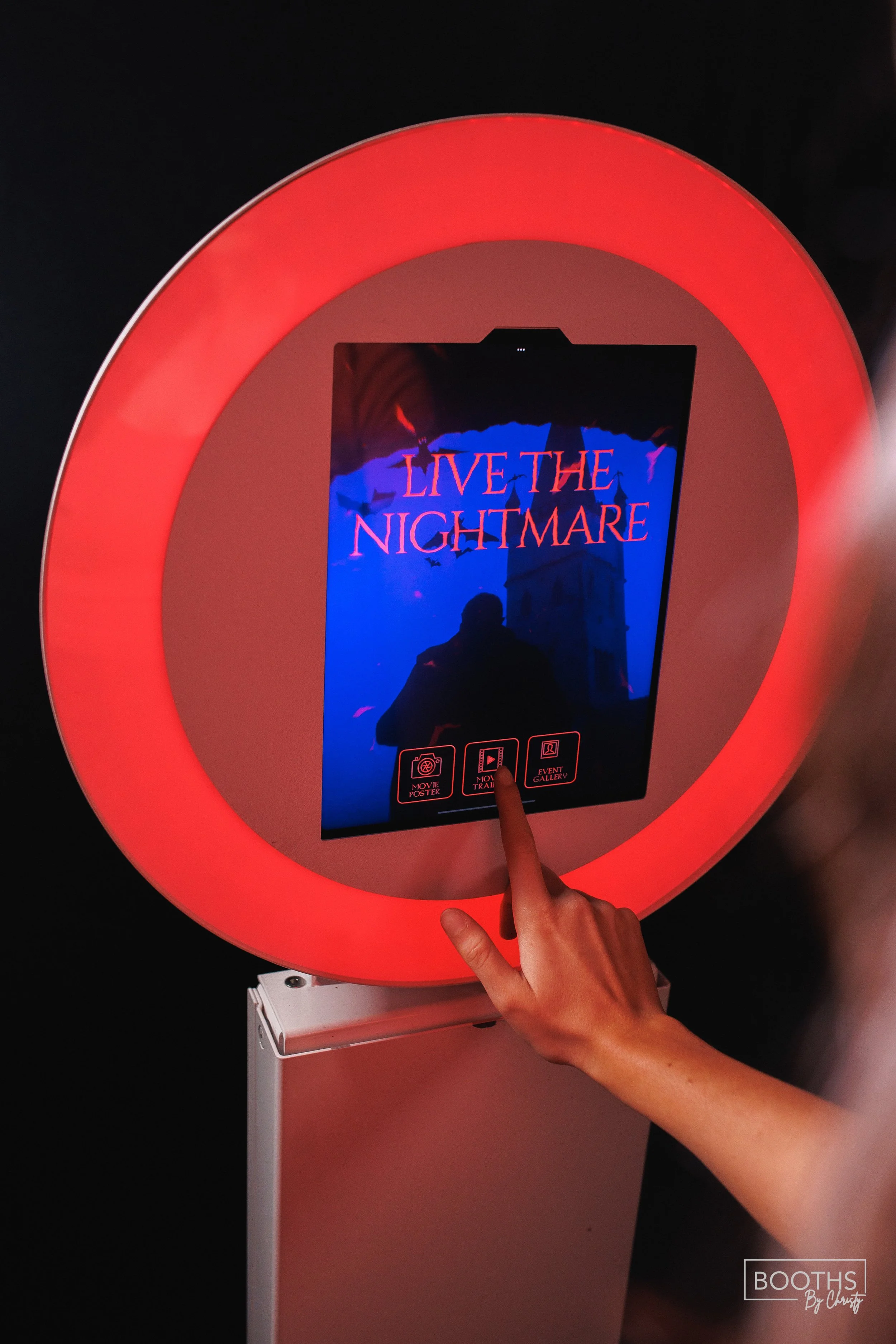 Touchscreen display with a red neon circular frame, showing the title 'LIVE THE NIGHTMARE' in red text, with icons for movie poster, movie trailer, and event gallery beneath. A person's hand is pointing at the screen.