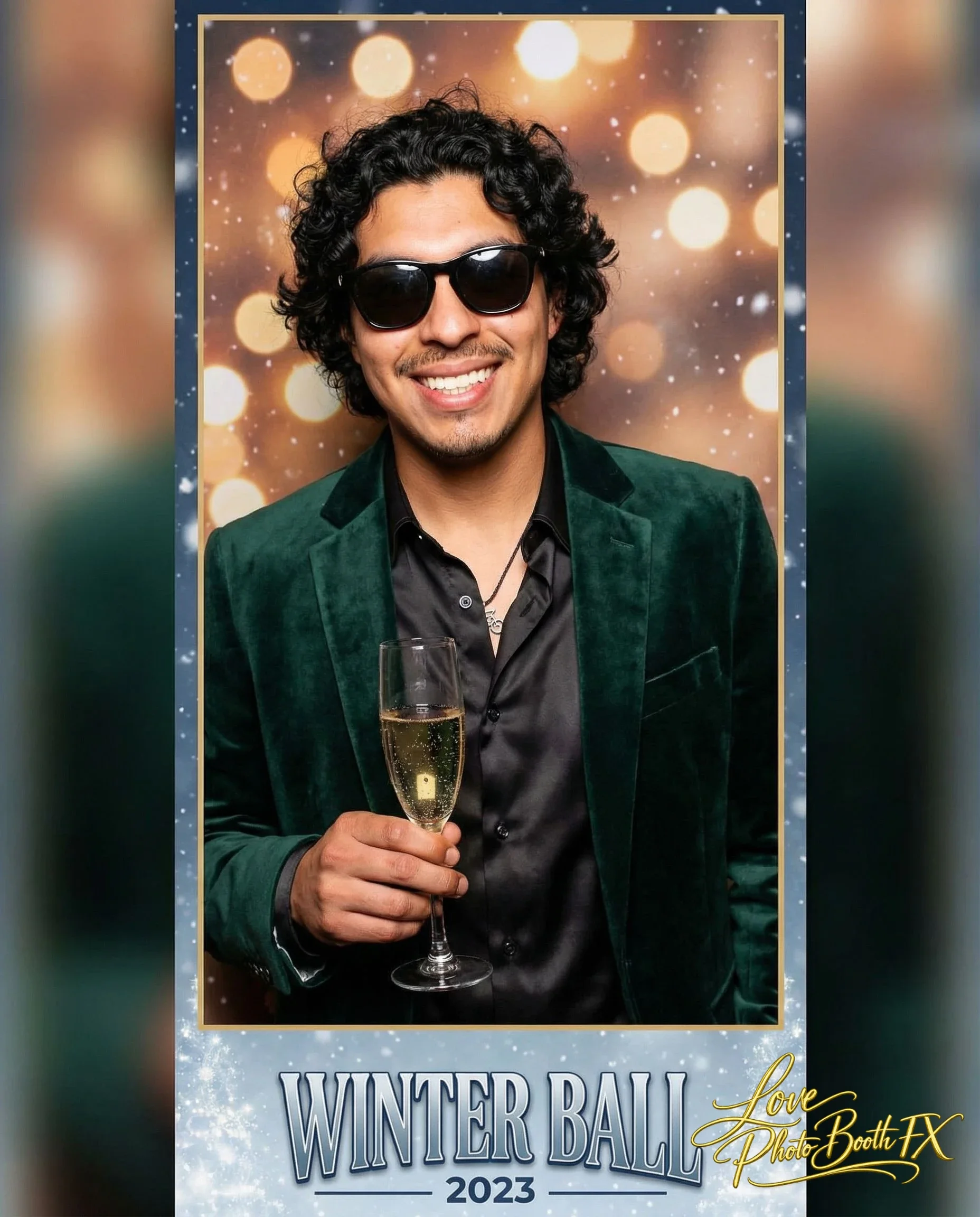 A man with curly black hair, wearing dark sunglasses and a green velvet blazer, smiles and holds a glass of champagne against a festive background with blurred golden lights. The photo is labeled 'Winter Ball 2023' from 'Love Photo Booth FX'.